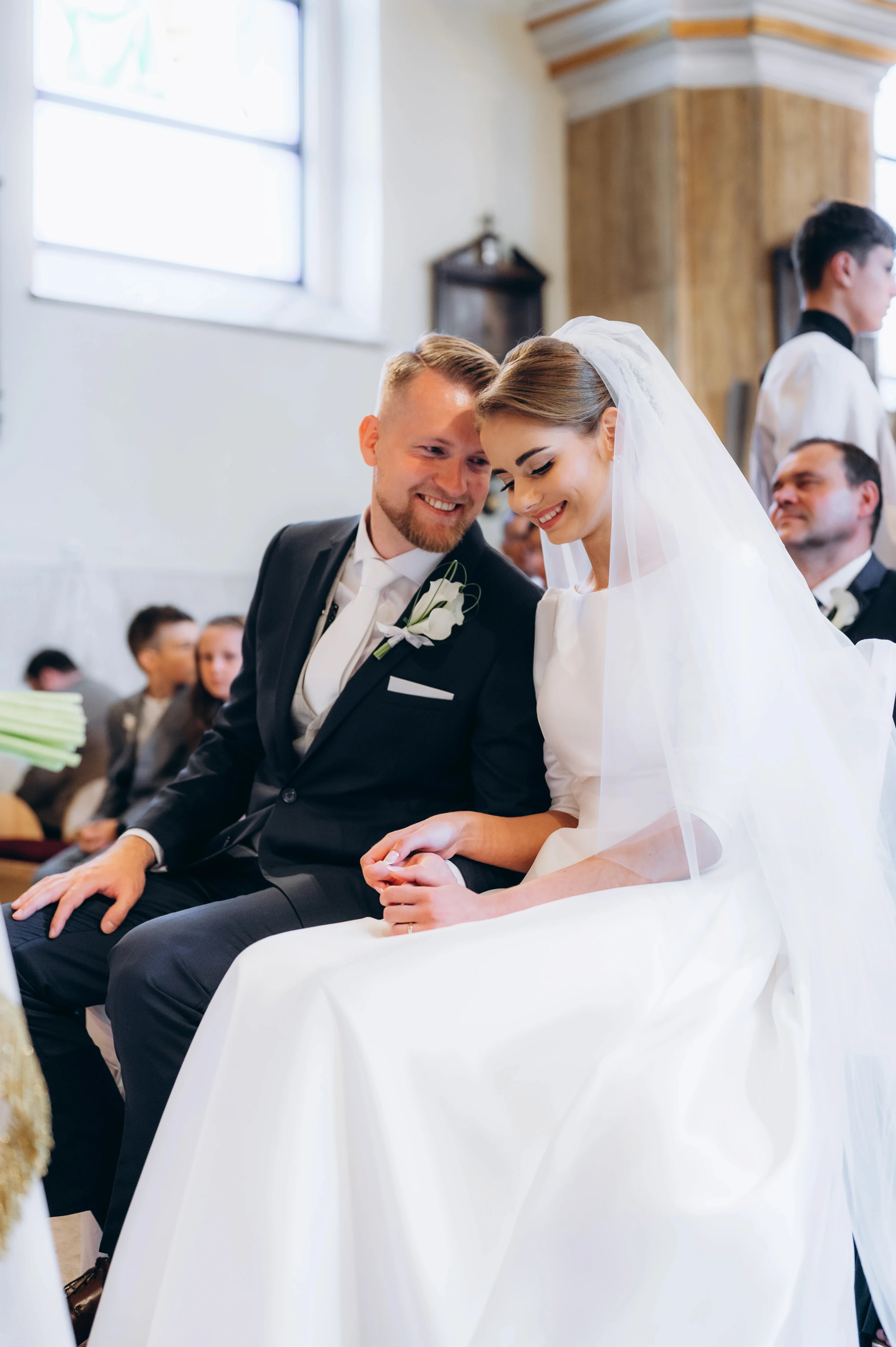 Ženích a nevesta so závojom sedia s hlavami pri sebe a usmievajú sa počas obradu v kostole.