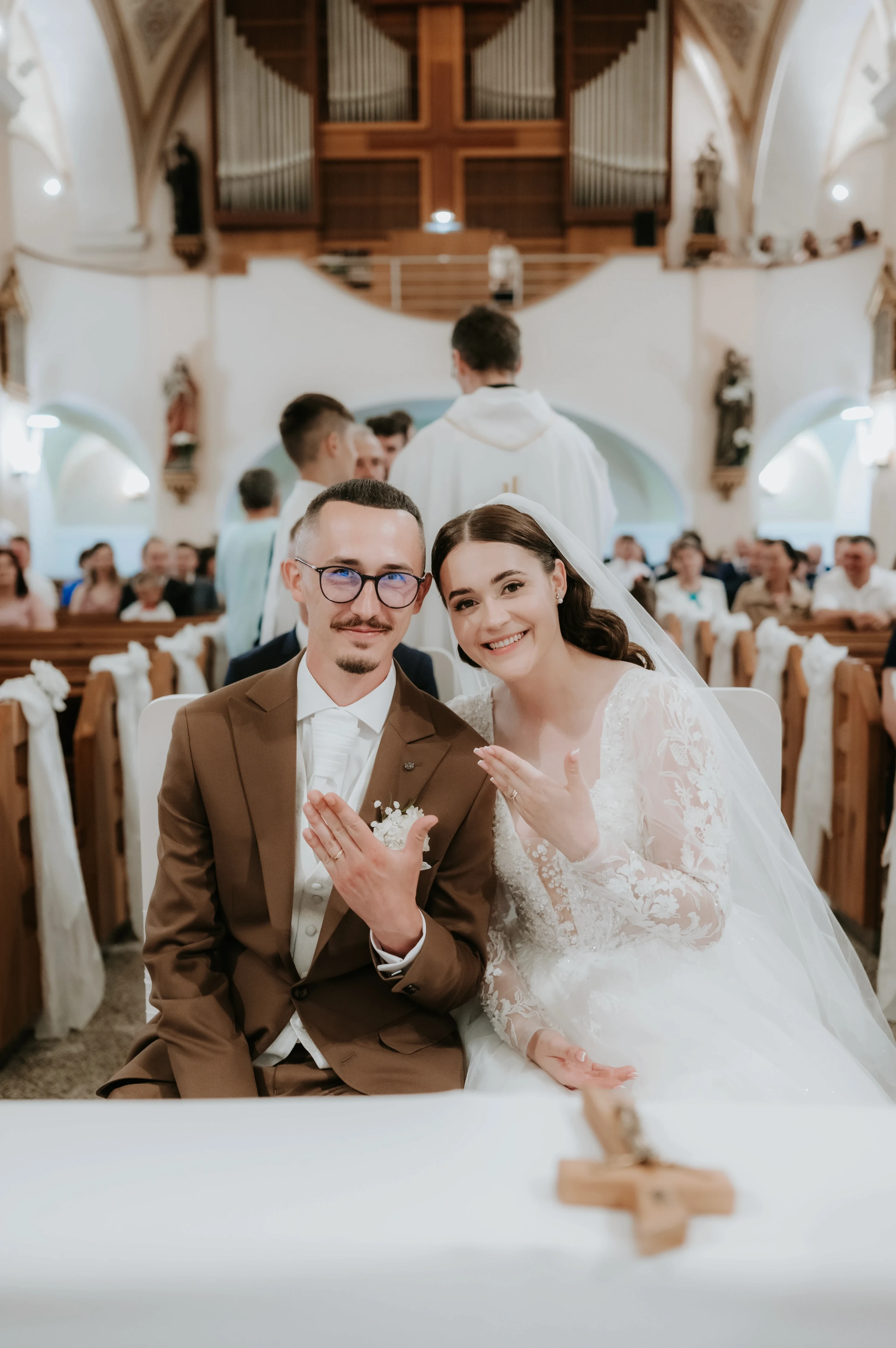 Ženích v hnedom obleku a nevesta v čipkových šatách sedia v kostolnej lavici a mávajú.