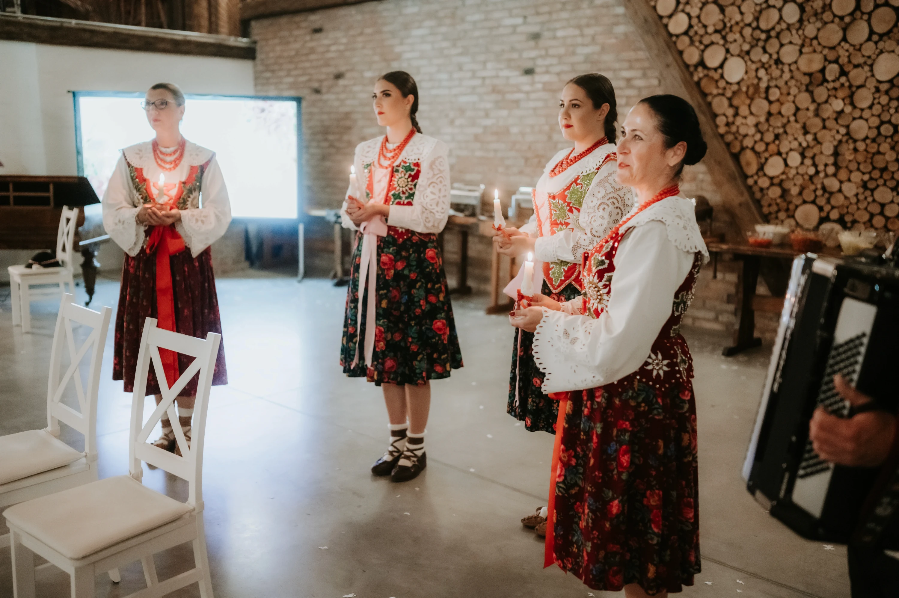 Štyri ženy v ľudovom kroji stoja so sviečkami, vpravo hrá harmonikár.