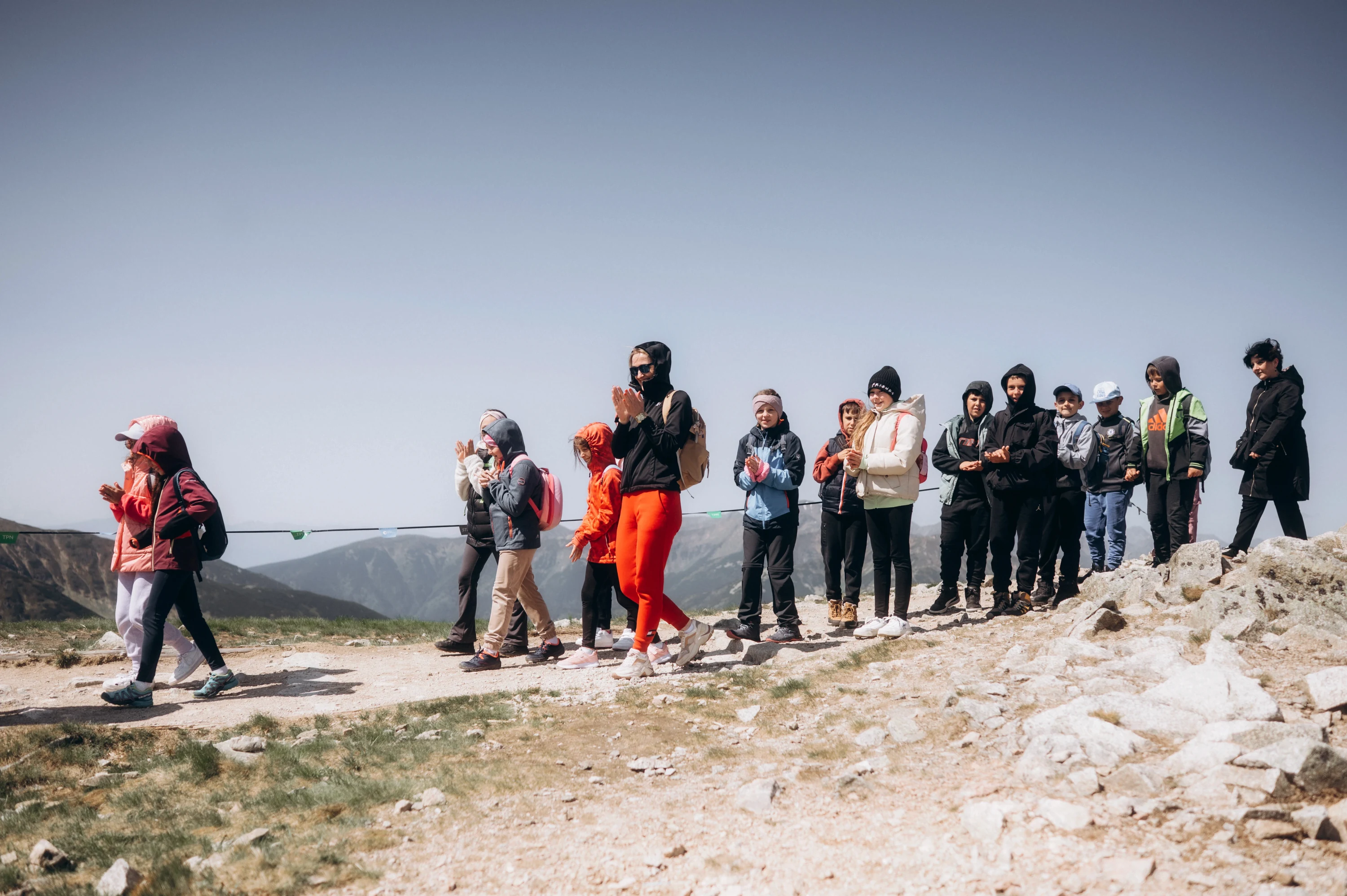 Skupina turistov v bundách kráča po kamenistom hrebeni hory pod jasnou oblohou.