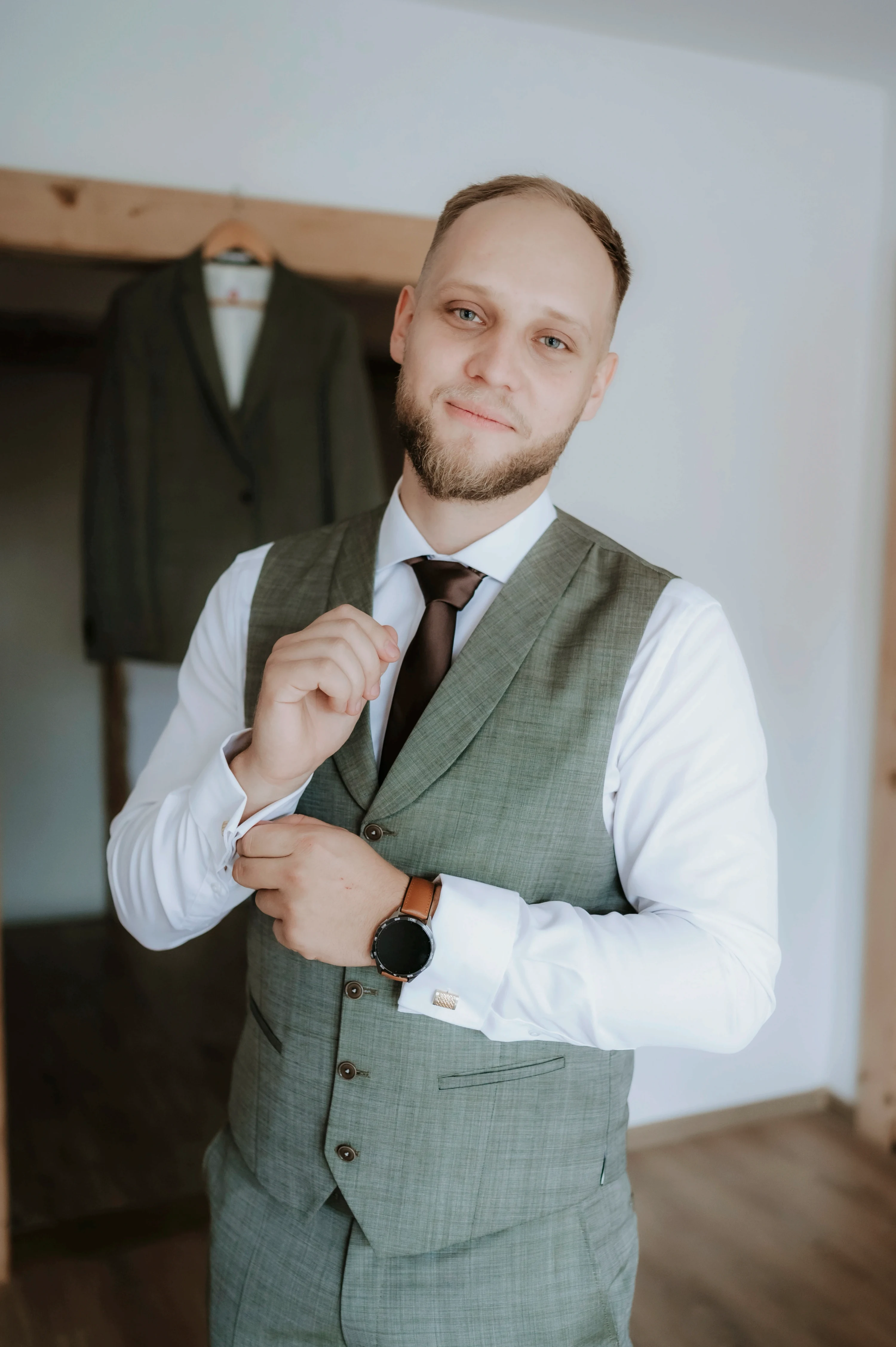 Ženích v sivej veste si upravuje manžetu, za ním visí na tráme sako.