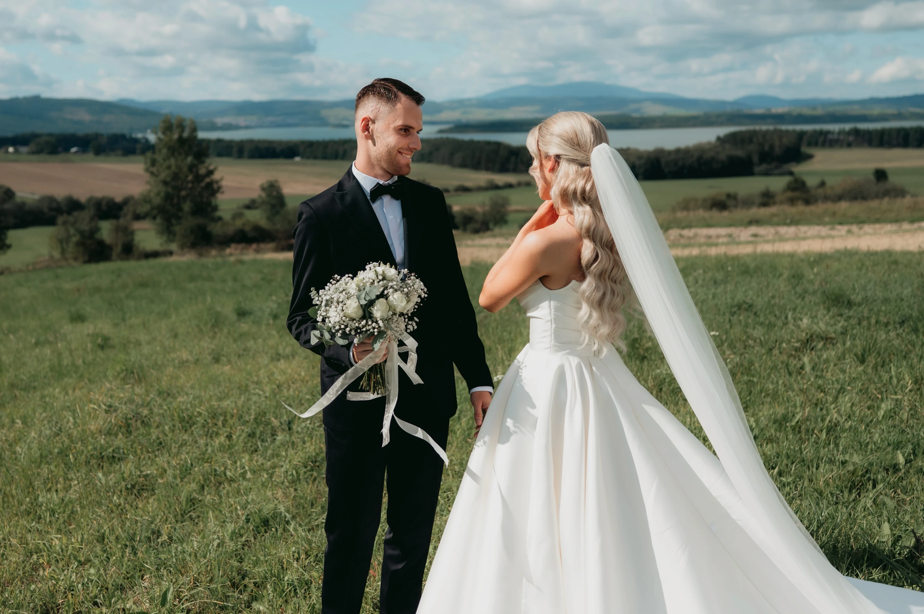 Ženích s kyticou stojí oproti neveste na lúke, v pozadí kopce a lesy.