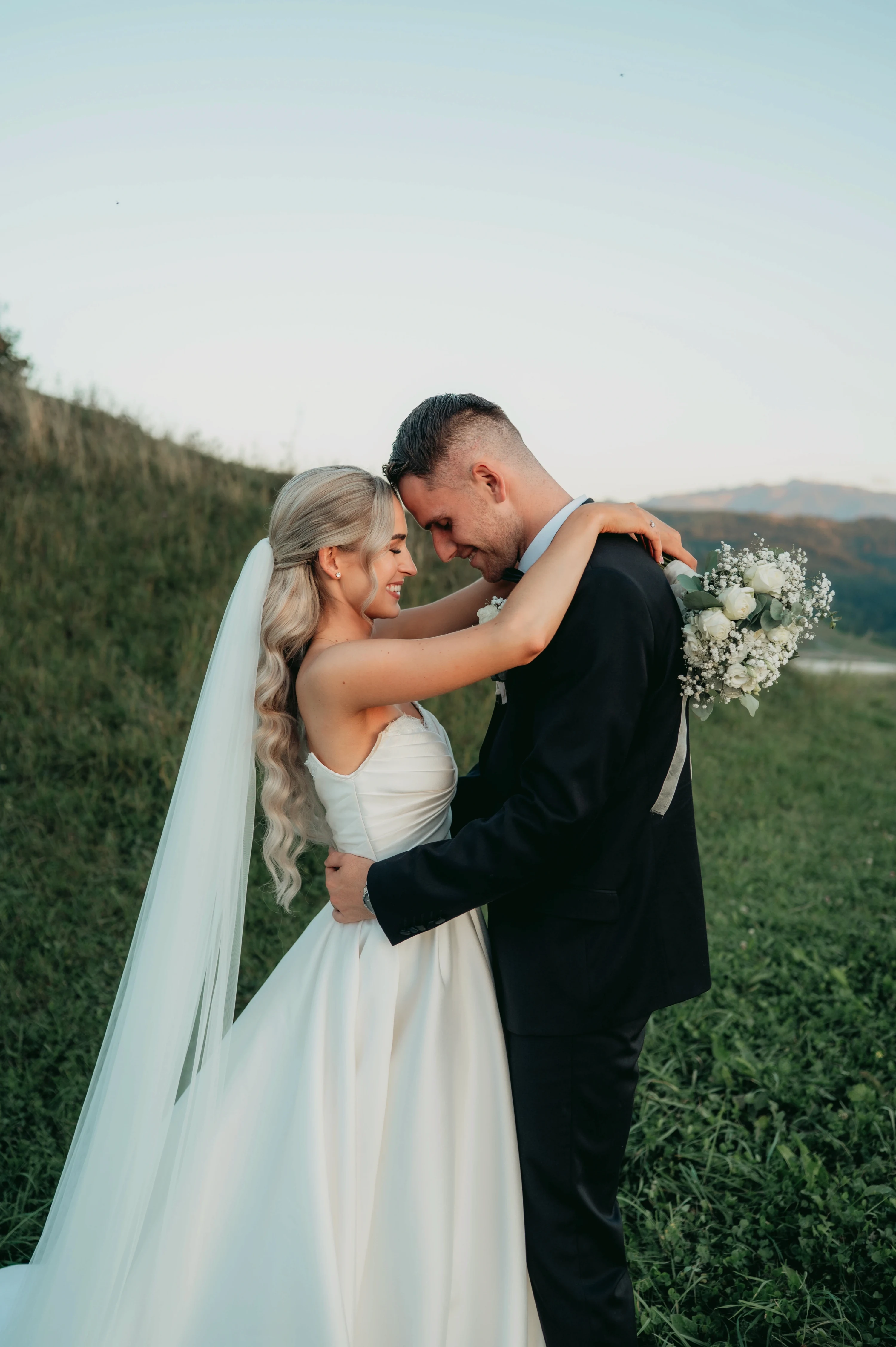 Katarína a Matej objíma svoju partnerku zozadu pri západe slnka.