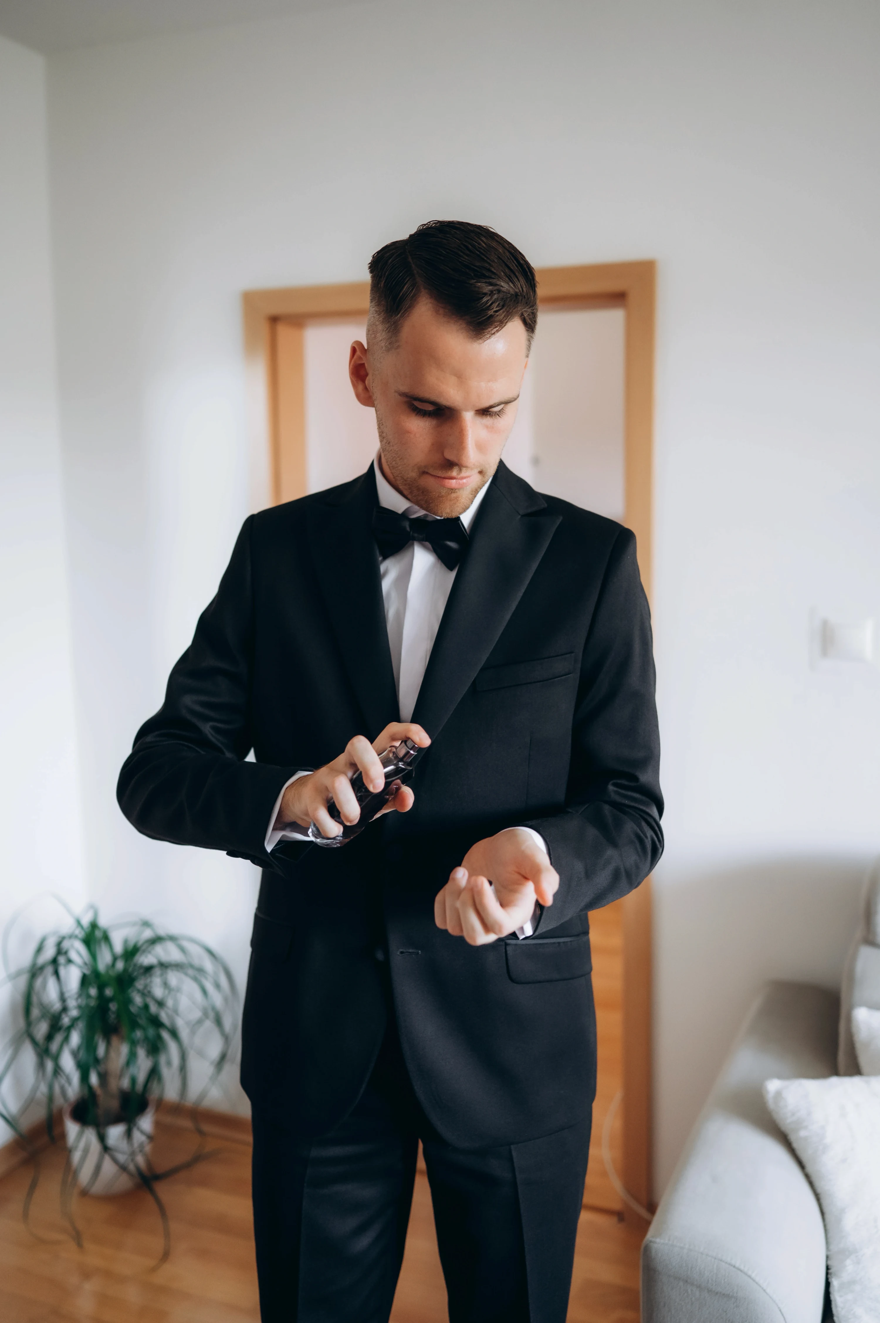 Ženích v čiernom smokingu s motýlikom si vo svetlej izbe upravuje manžetu na ľavej ruke.
