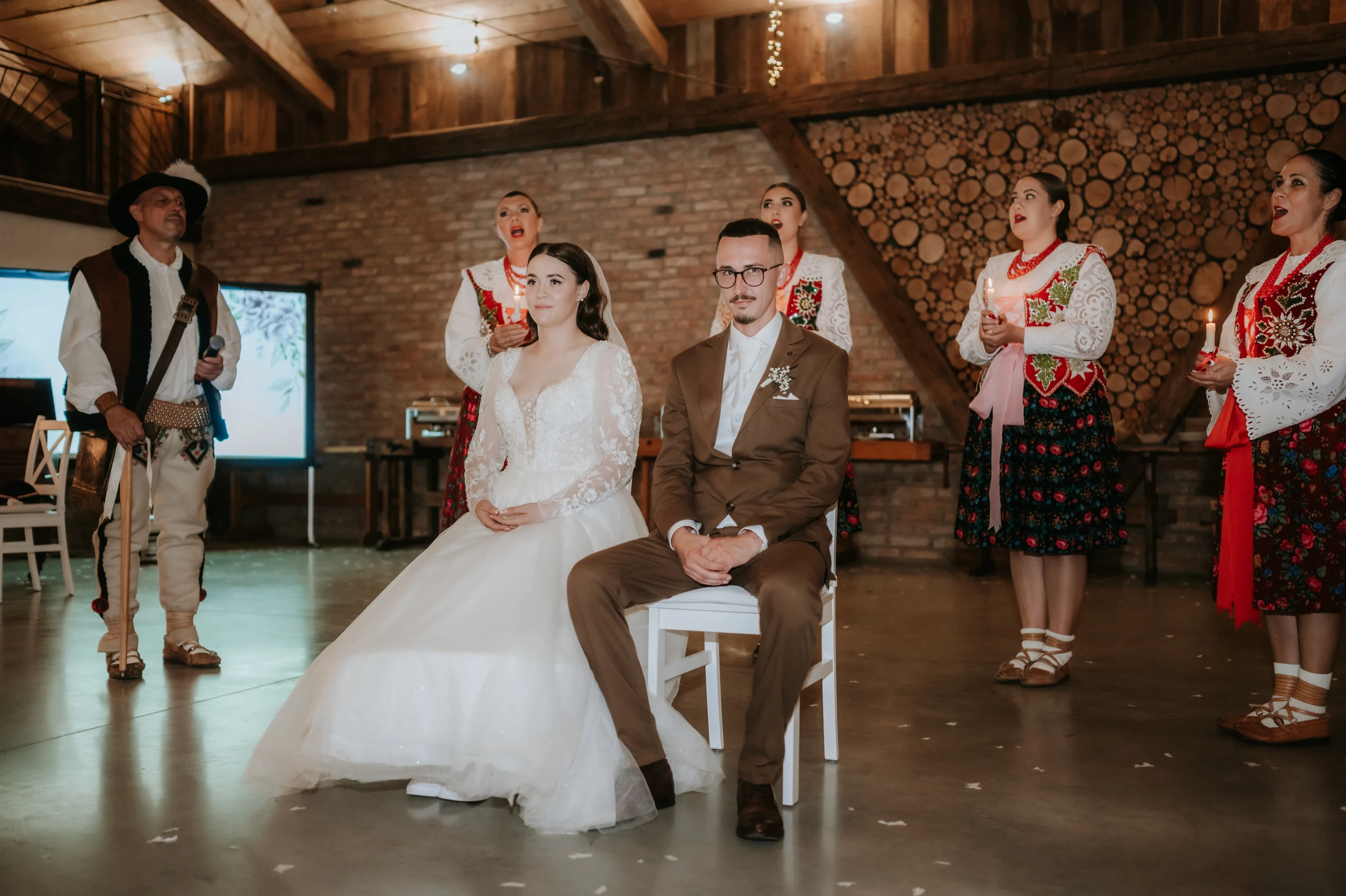 Novomanželia sedia na stoličkách v stodole, okolo nich spievajú ženy a muž v ľudovom kroji.