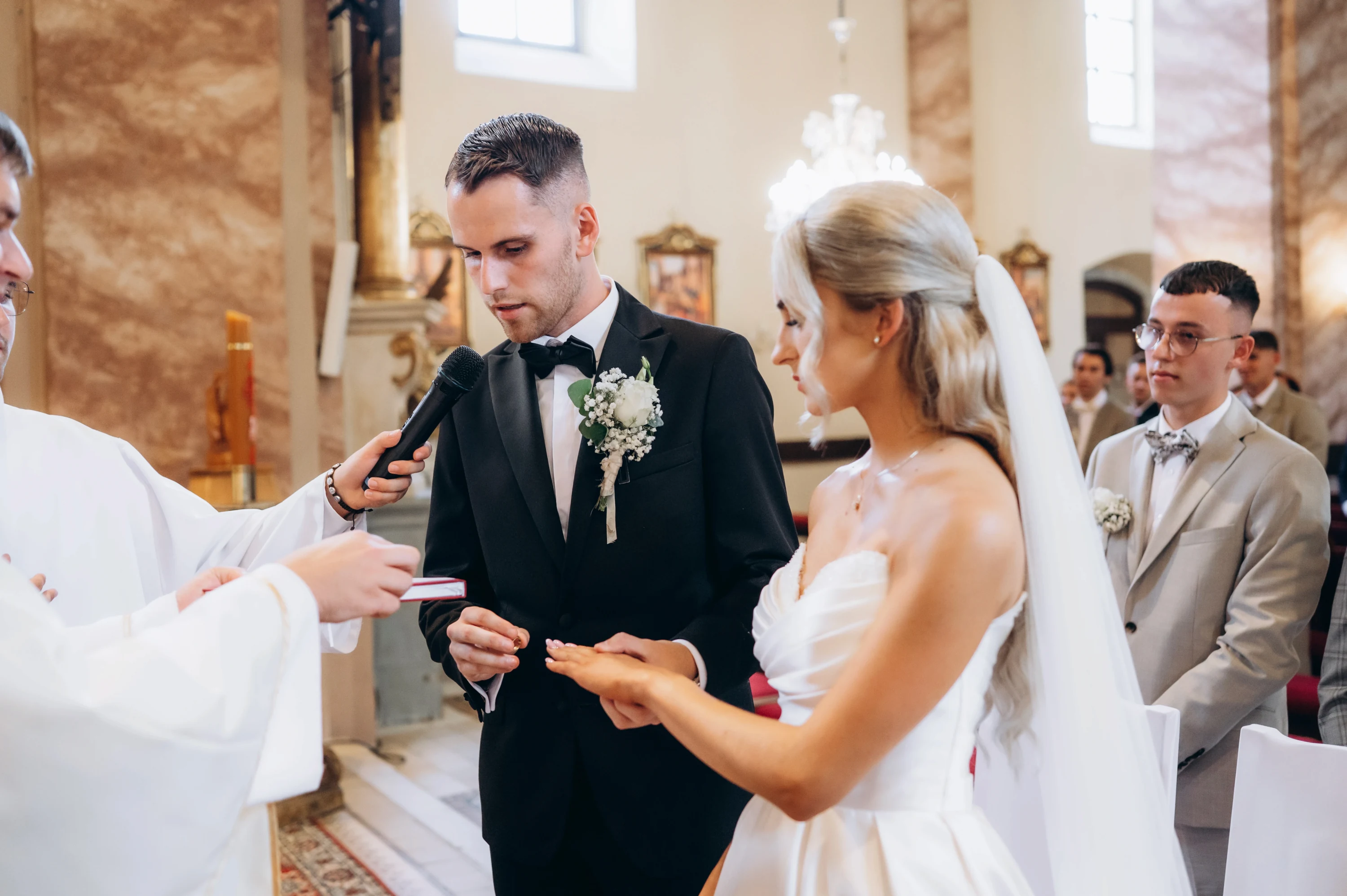 Kňaz s mikrofónom stojí pred ženíchom a nevestou počas výmeny obrúčok v kostole.
