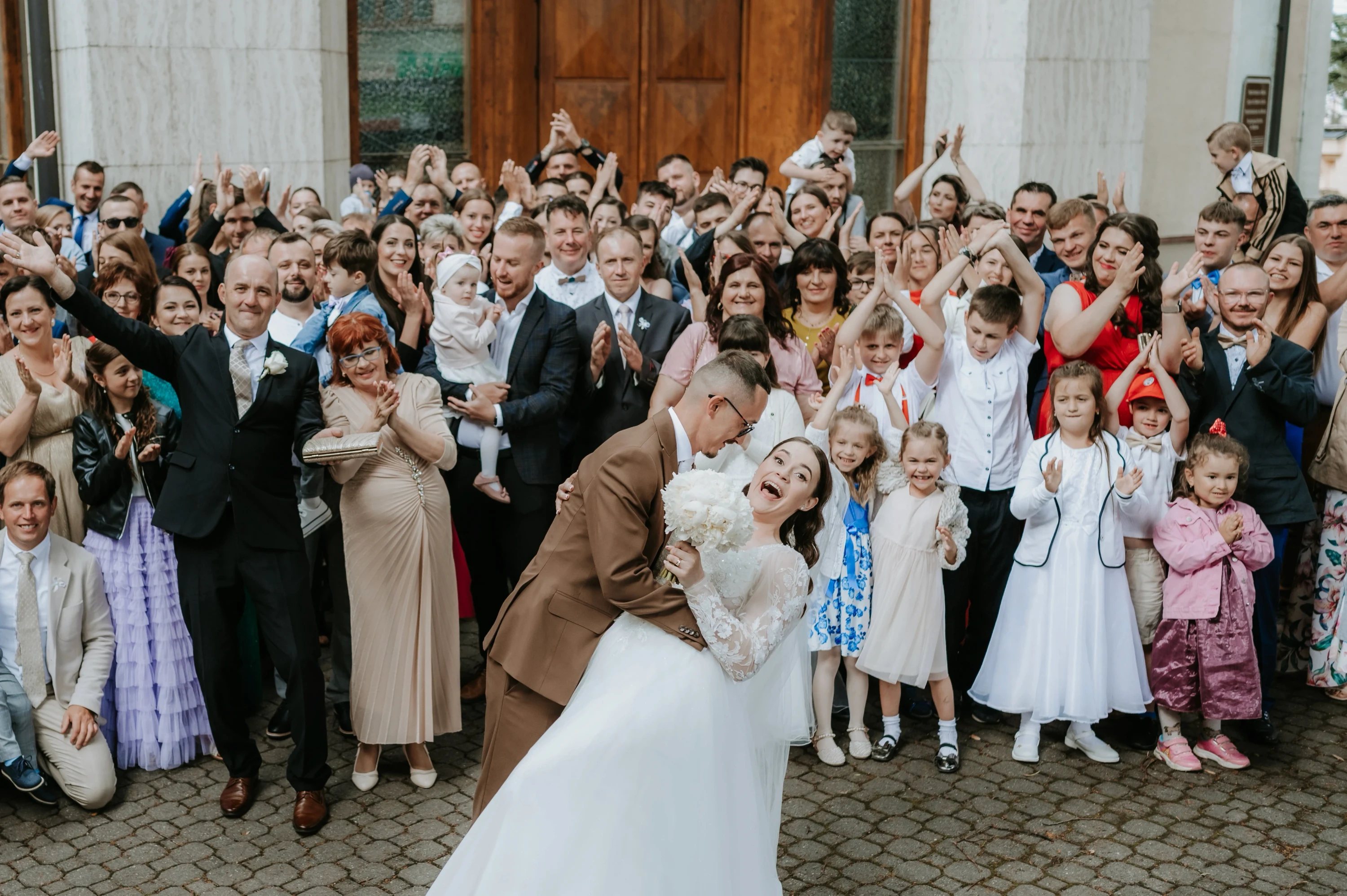 Ženích zakláňa nevestu k bozku pred jasajúcim zástupom hostí pred budovou.