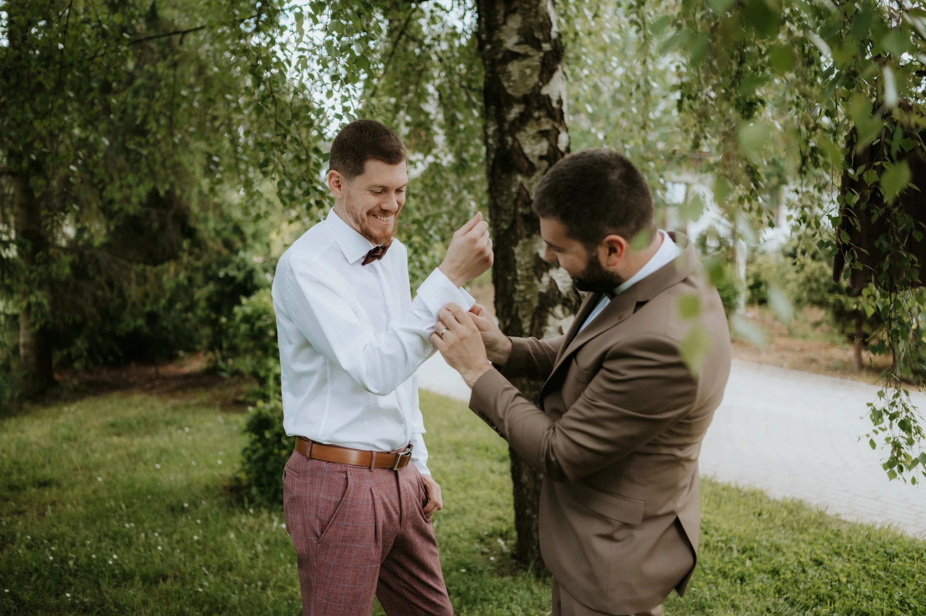 Muž v hnedom obleku pomáha druhému s manžetou košele v záhrade pod brezami.