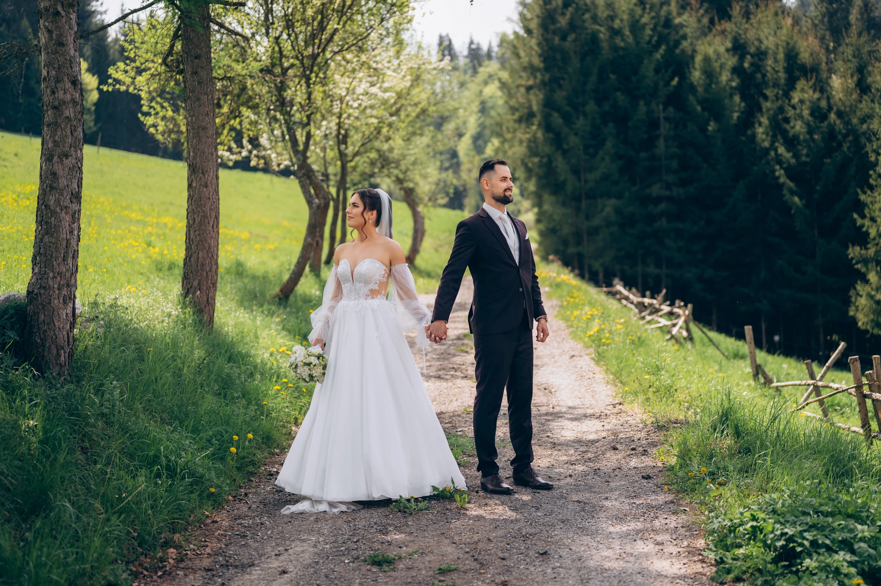 Novomanželia sa držia za ruky na lesnej ceste obklopenej prírodou.