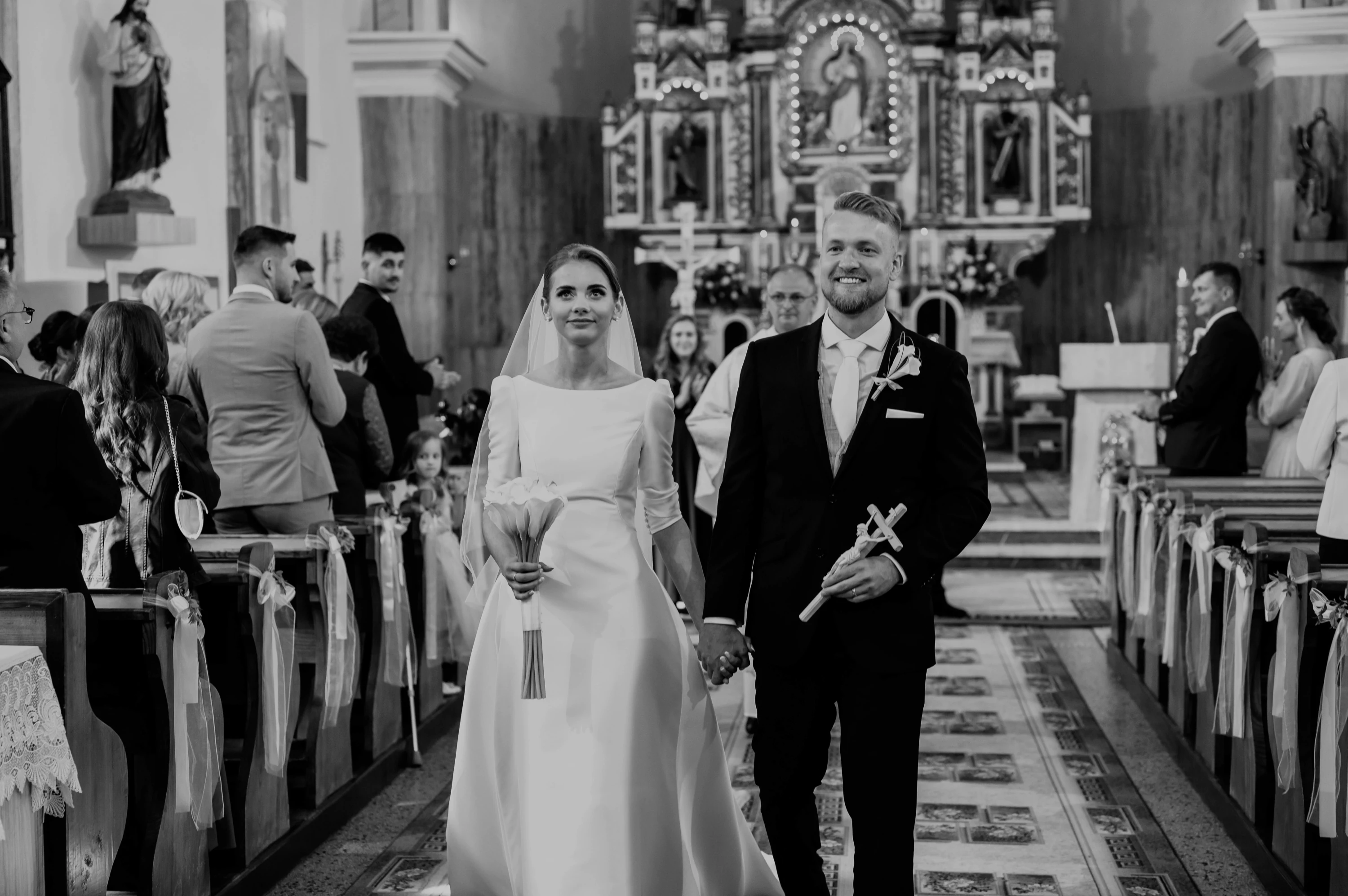 Čiernobiely záber: novomanželia sa držia za ruky a kráčajú uličkou zaplneného kostola.