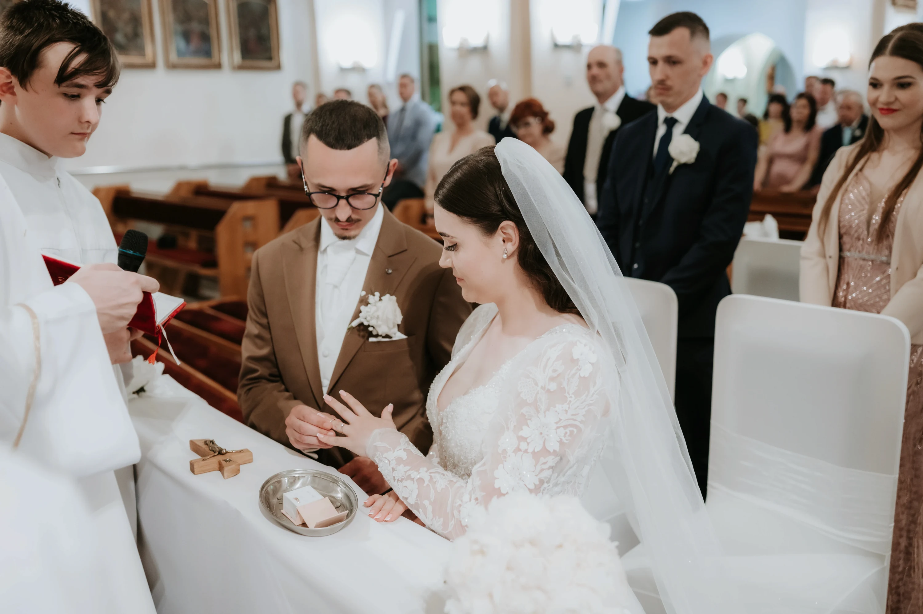 Ženích navlieka neveste obrúčku pri oltári, miništrant vedľa nich drží mikrofón.