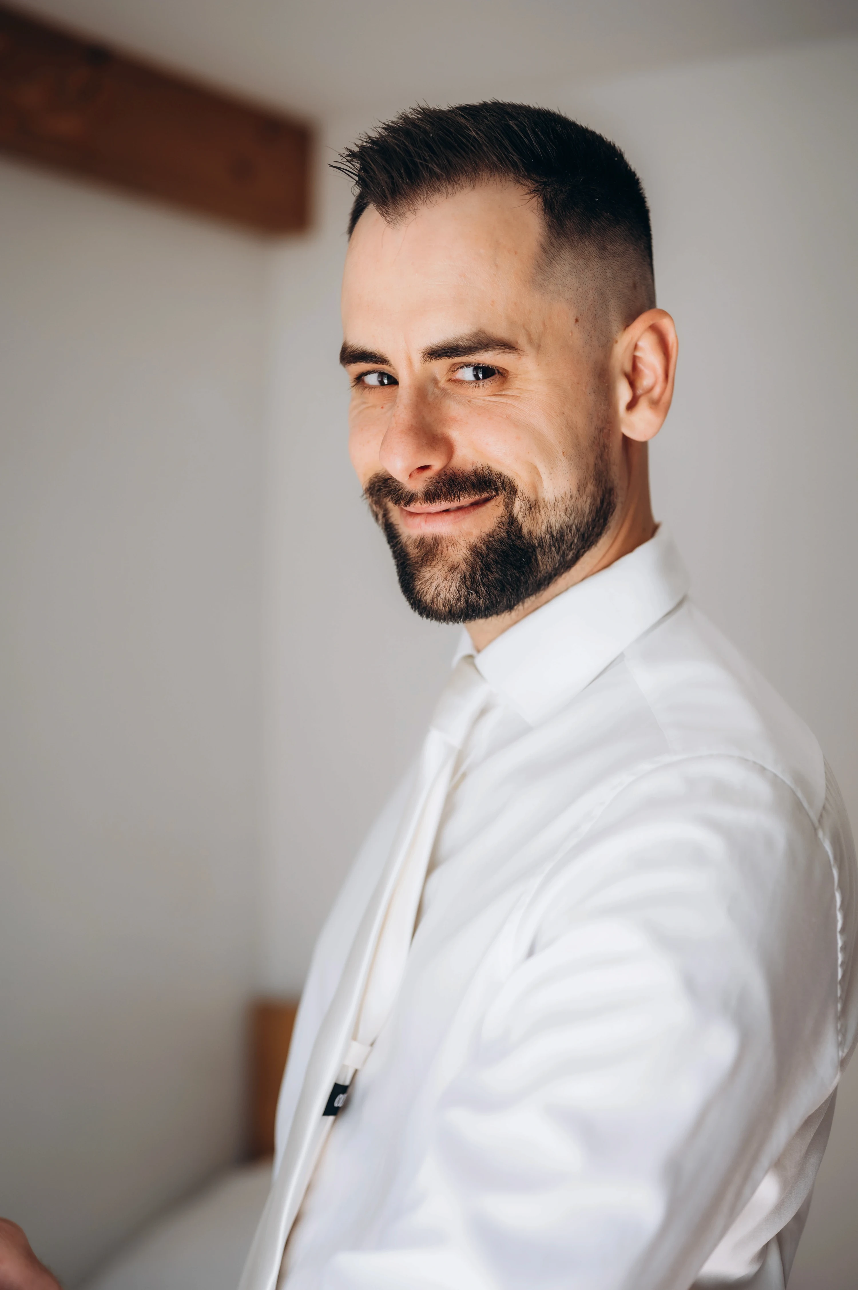 Ženích v bielej košeli sa cez plece usmieva do objektívu počas príprav vo svetlej izbe.