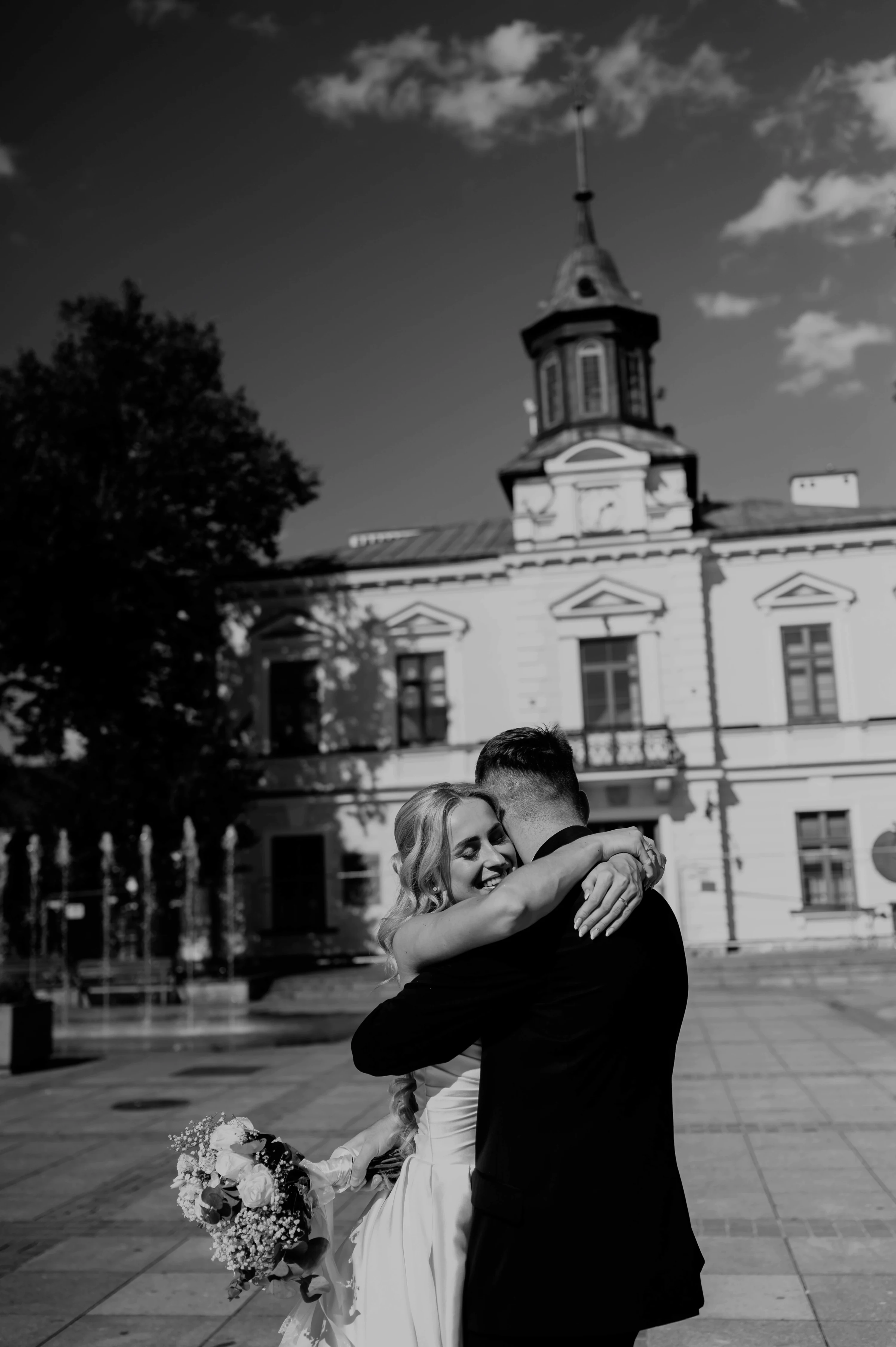 Čiernobiely záber: novomanželia sa objímajú na dláždenom priestranstve pred historickou budovou.