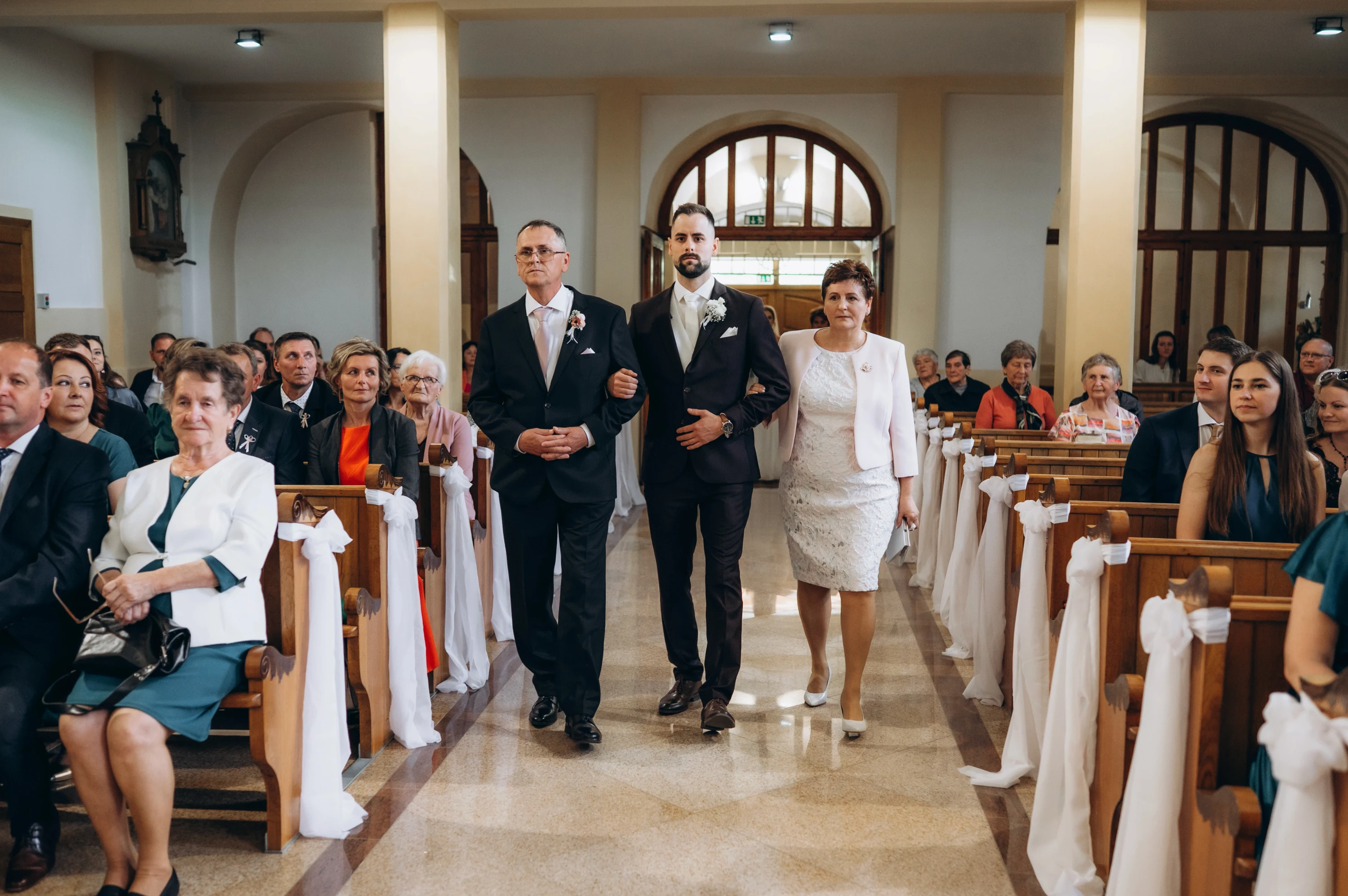 Ženích kráča kostolnou uličkou s rodičmi medzi hosťami.