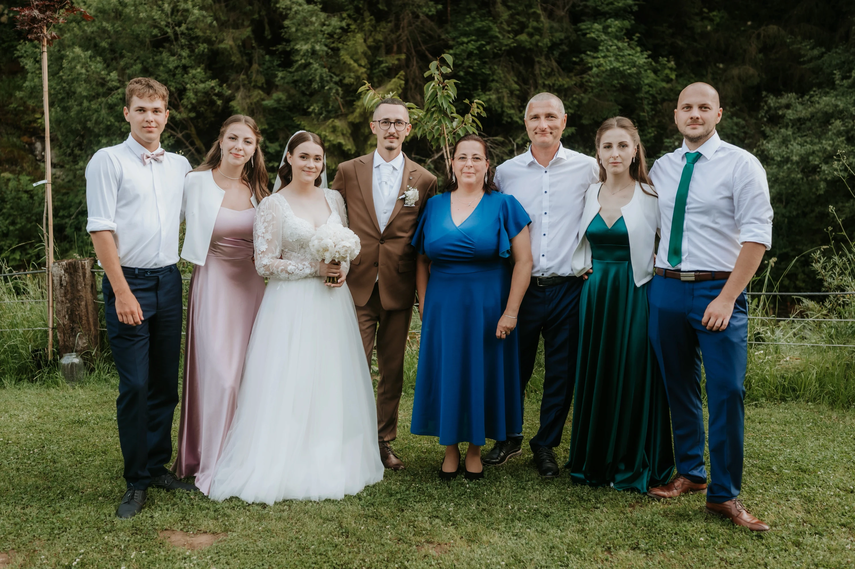 Skupinový portrét: novomanželia stoja s rodinou v tráve pred stromami.