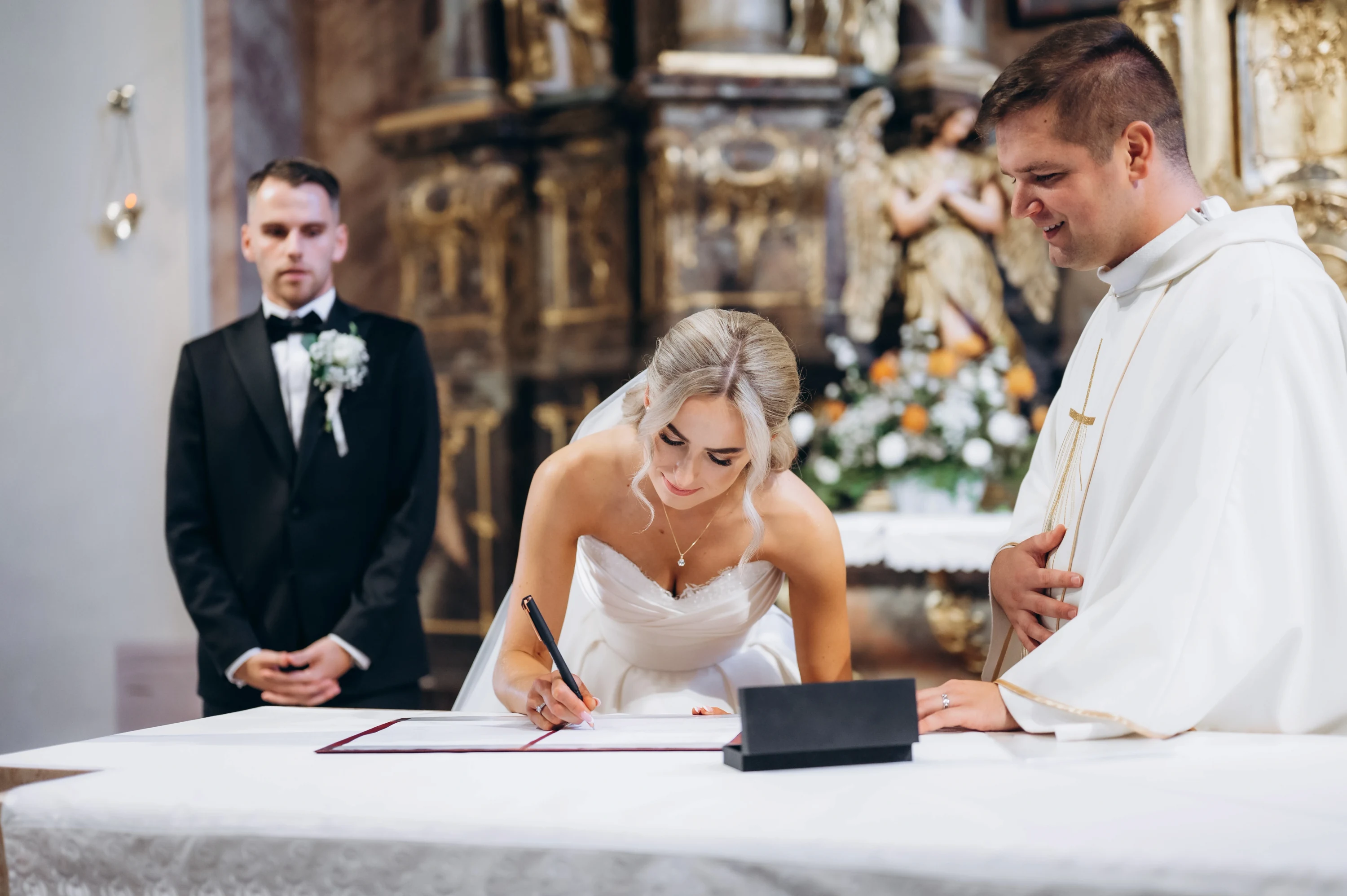 Nevesta podpisuje sobášny zápis, kňaz a ženích sa prizerajú.
