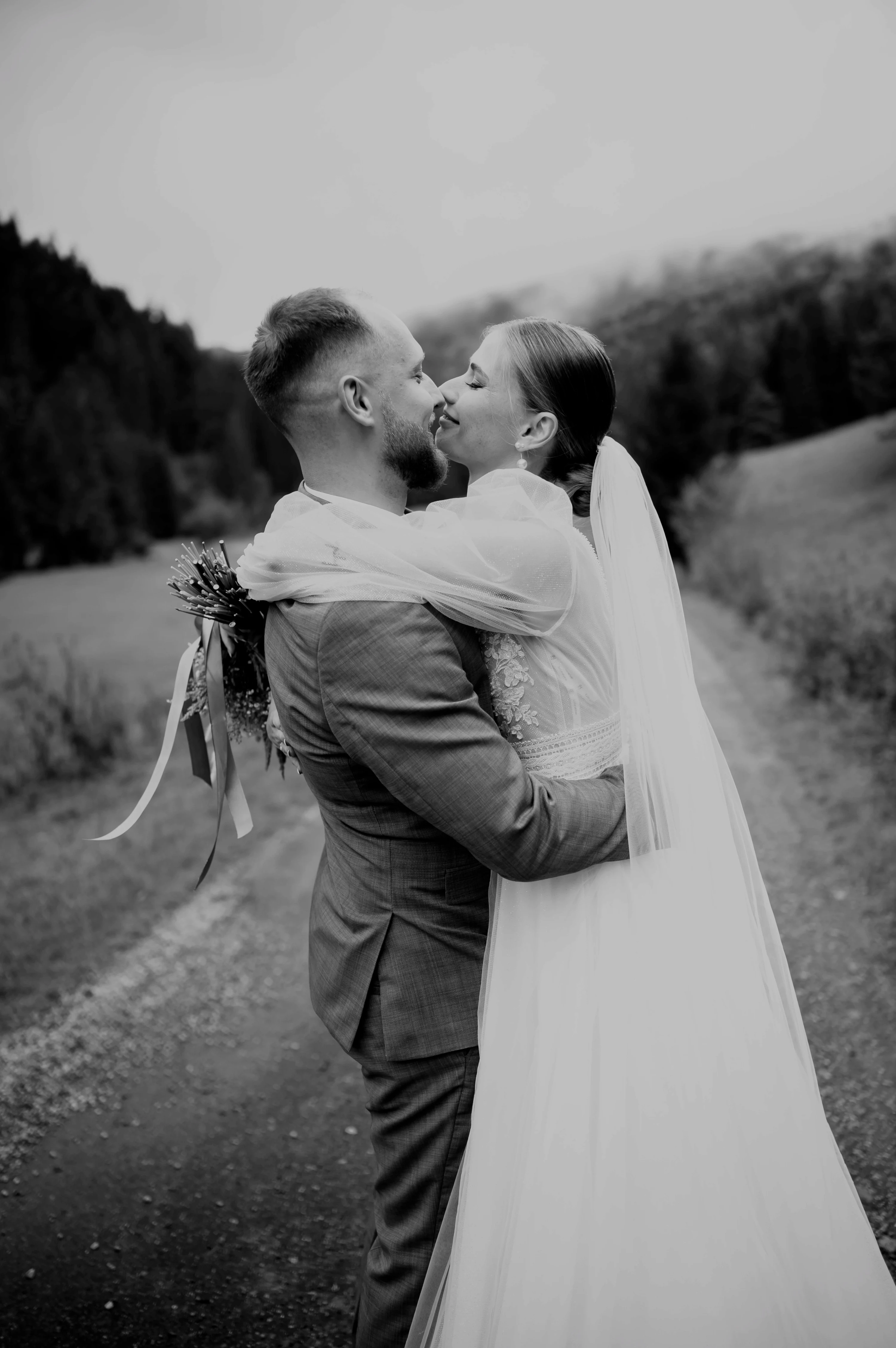 Čiernobiely záber: novomanželia sa bozkávajú na poľnej ceste medzi lúkami.