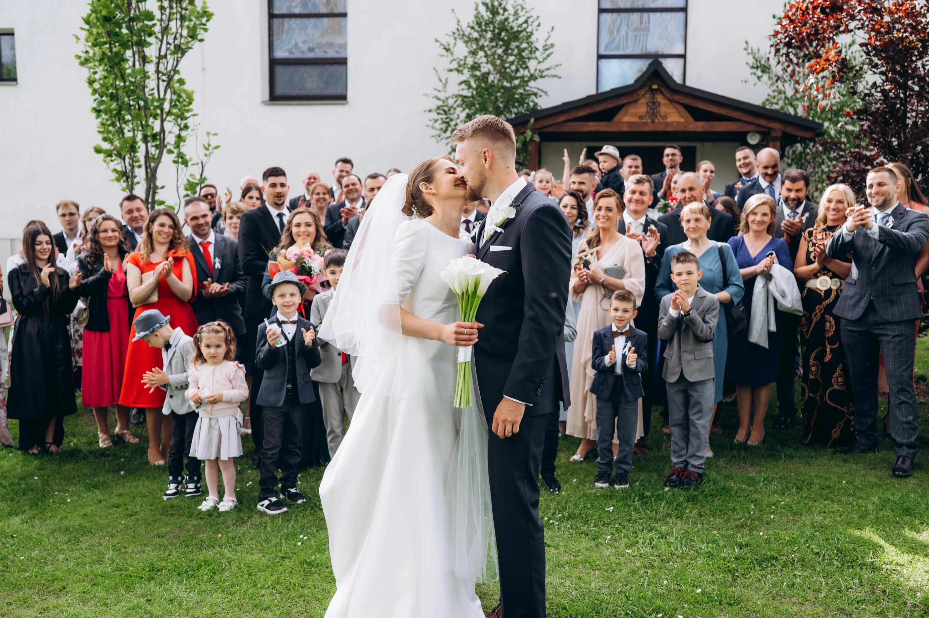 Novomanželia sa bozkávajú vonku s veľkou skupinou radostných hostí.