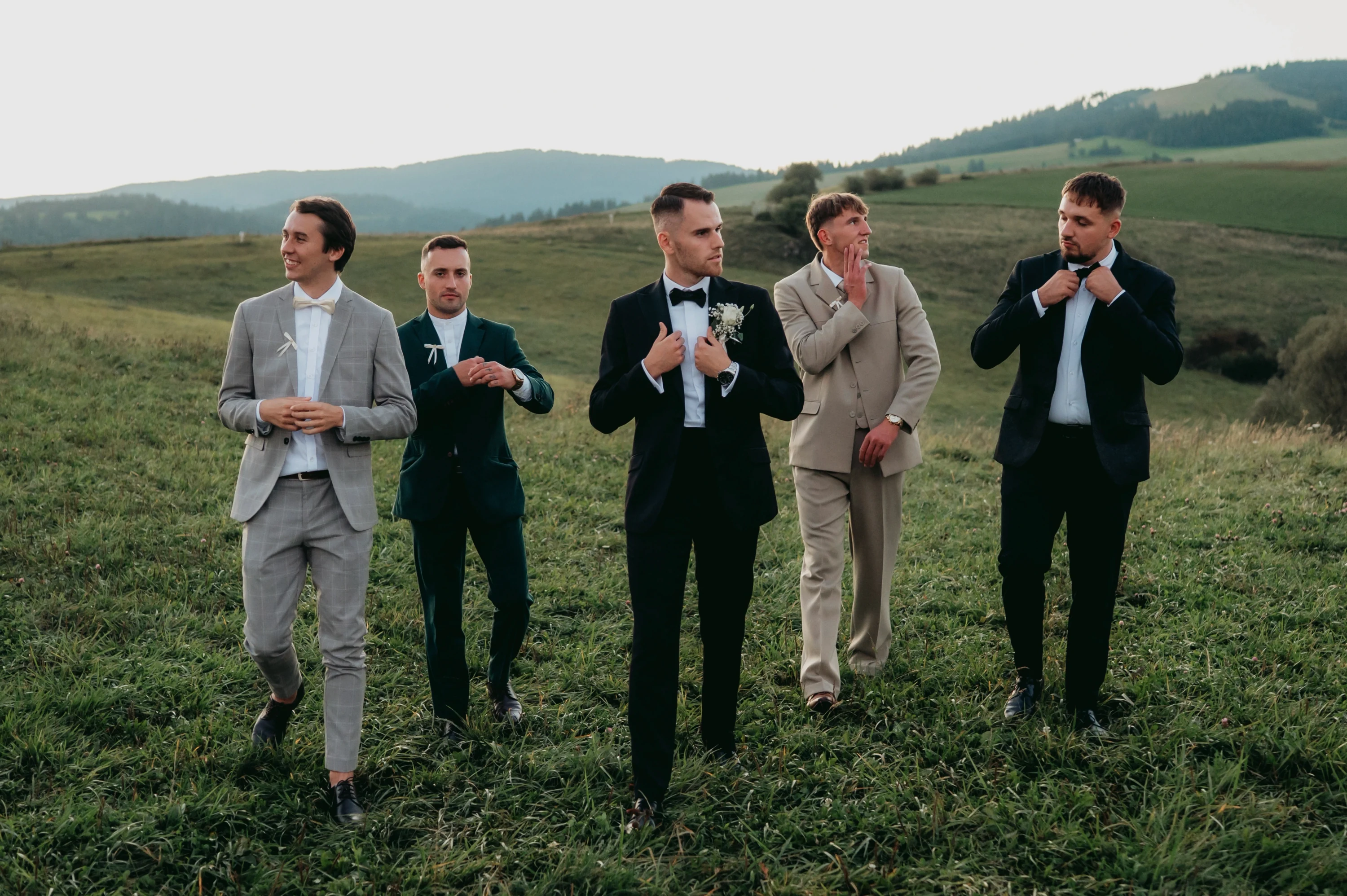 Piati muži v oblekoch kráčajú po lúke a upravujú si motýliky pred obradom.