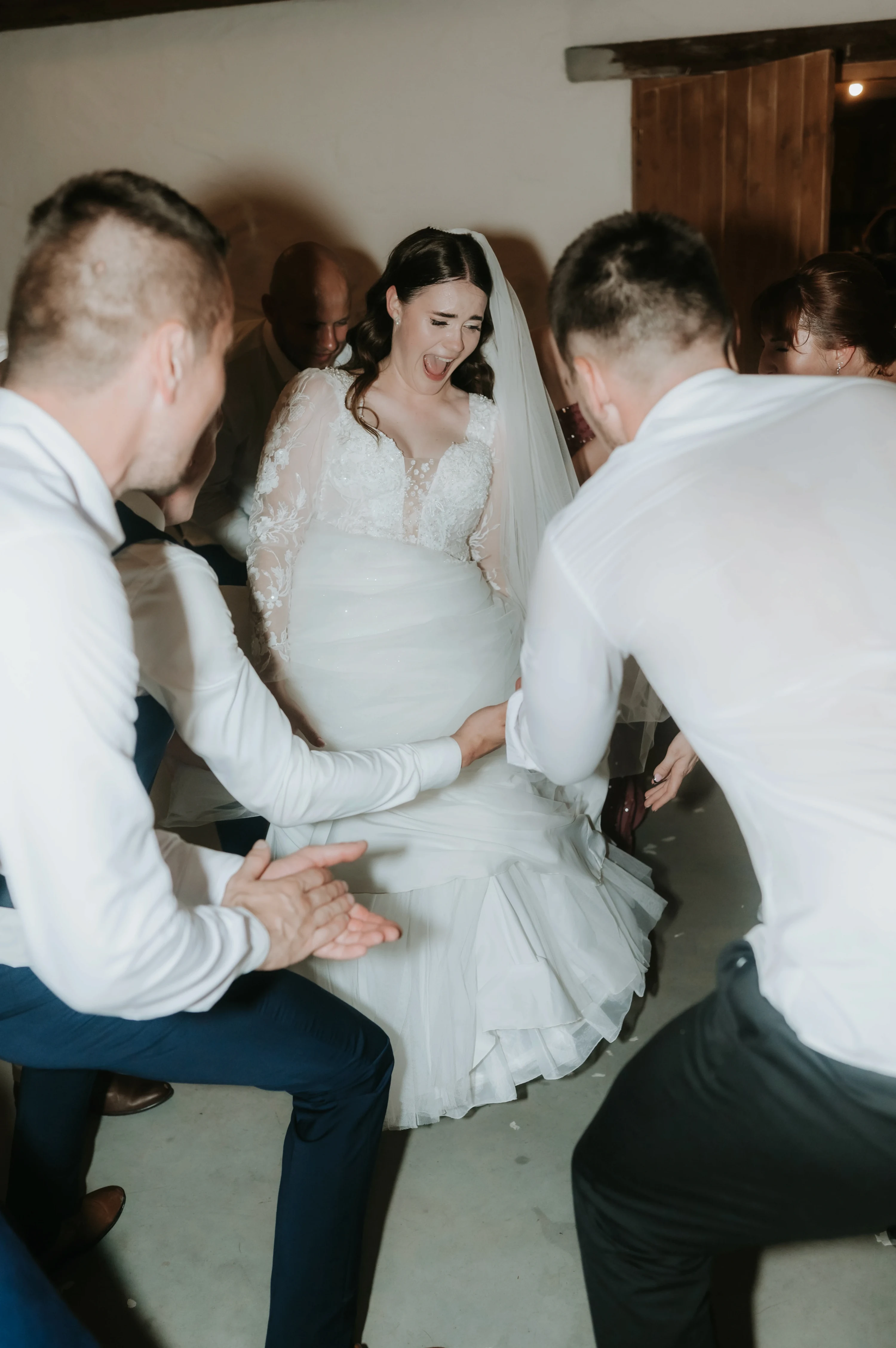 Smejúca sa nevesta sedí na stoličke, dvaja muži v bielych košeliach sa skláňajú okolo nej.