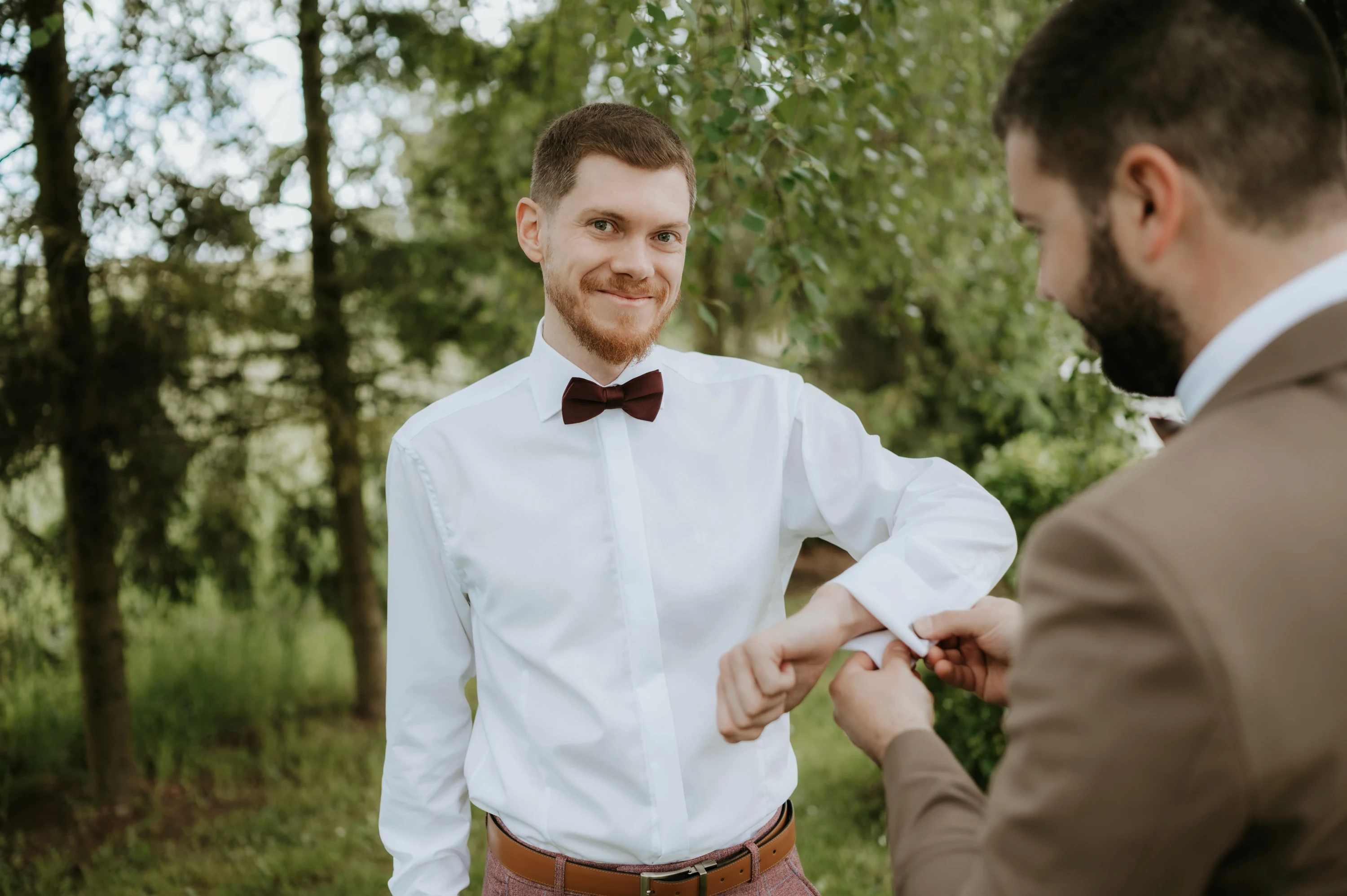 Priateľ v hnedom obleku zapína ženíchovi manžetu, ženích sa usmieva do objektívu.