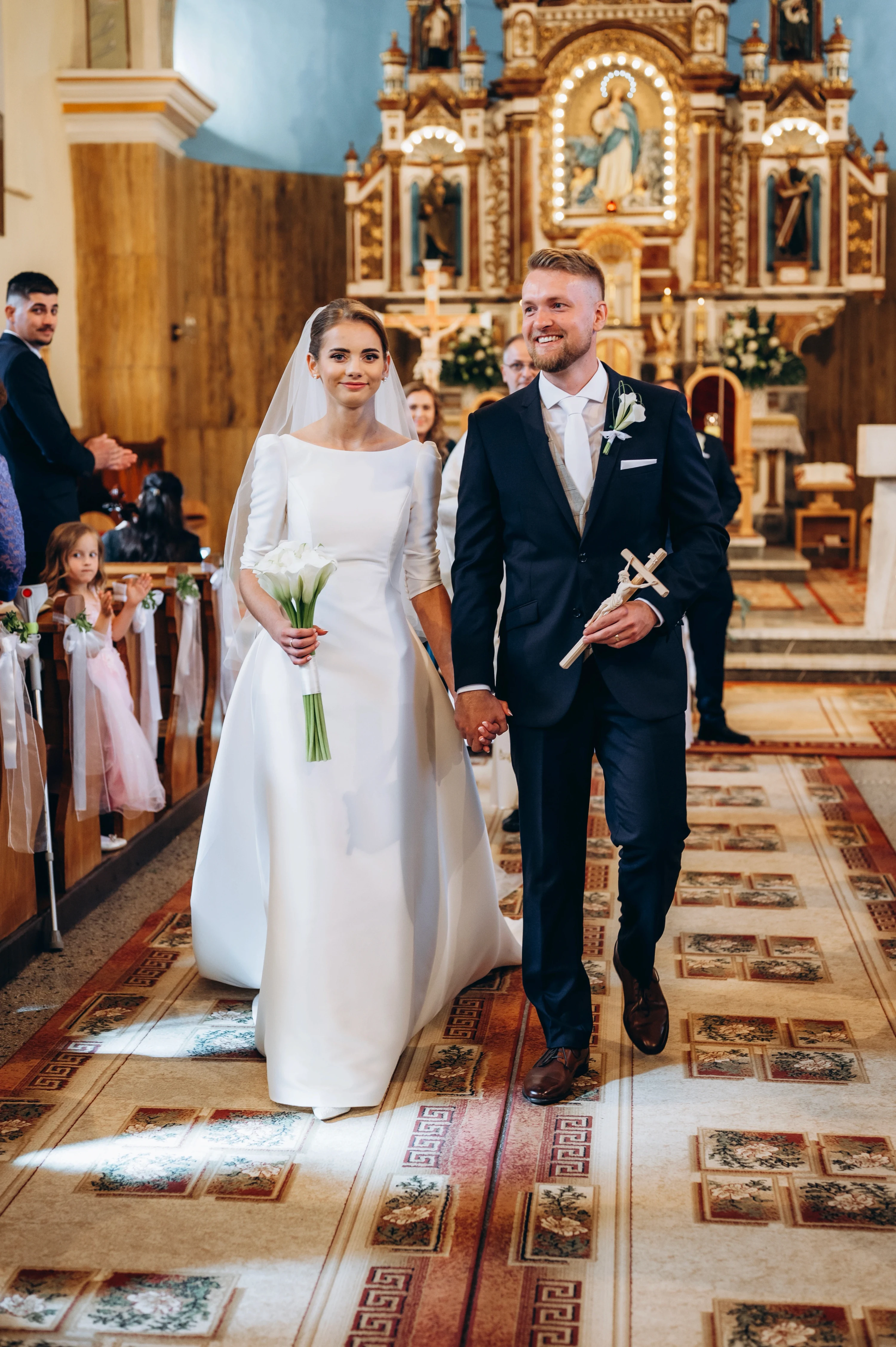 Novomanželia kráčajú do kostola so spojenými rukami počas obradu.