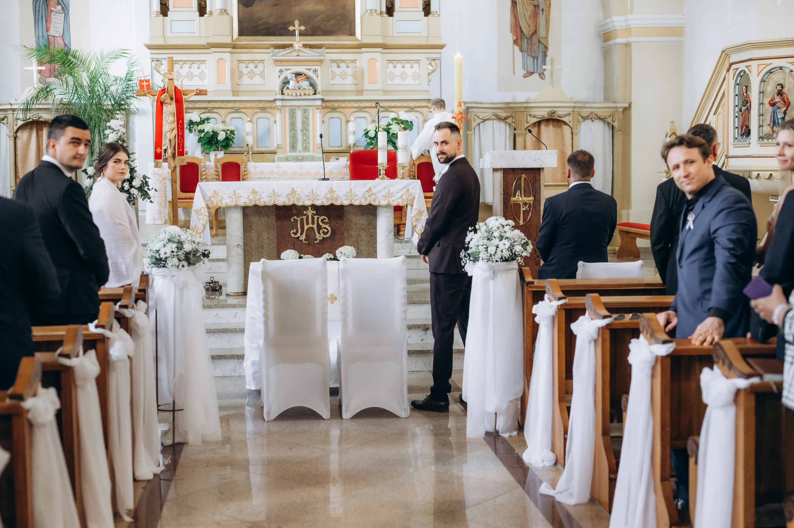 Ženích čaká pri oltári v kostole, hostia stoja v laviciach zdobených bielymi mašľami.