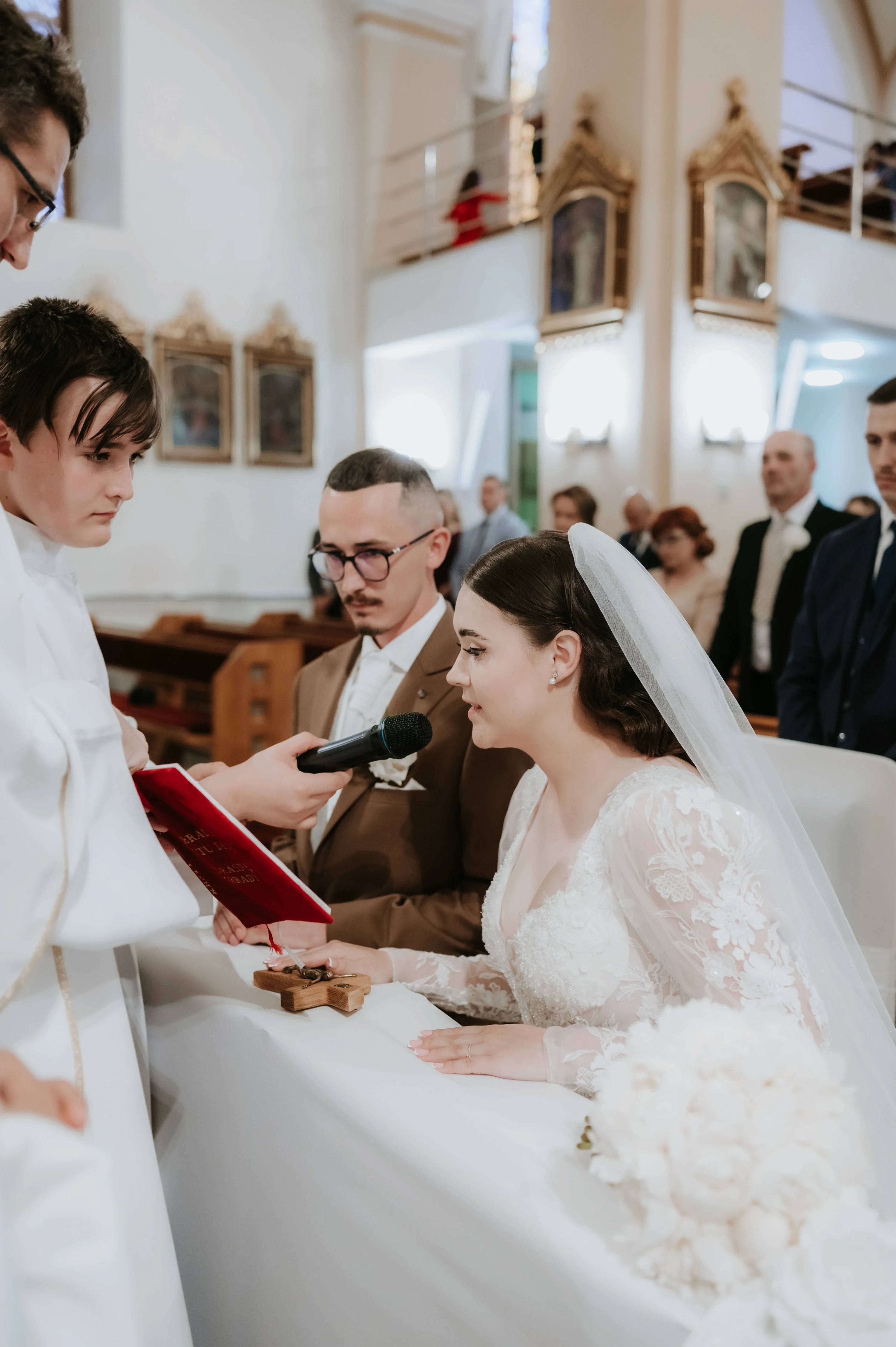 Kňaz s mikrofónom a červenou knihou stojí pred nevestou a ženíchom pri oltári.