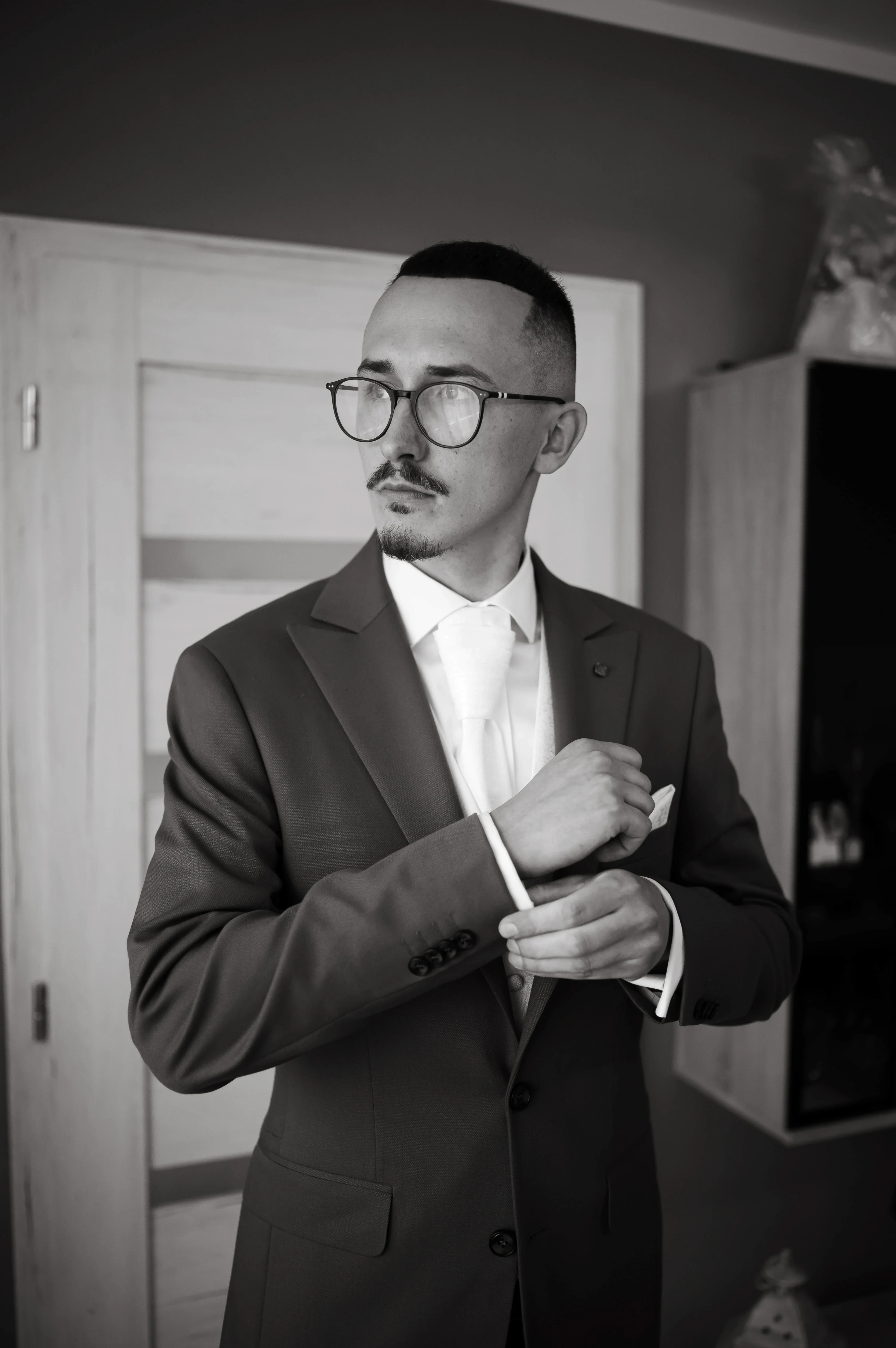 Čiernobiely záber: ženích v okuliaroch si v izbe upravuje manžetu bielej košele.