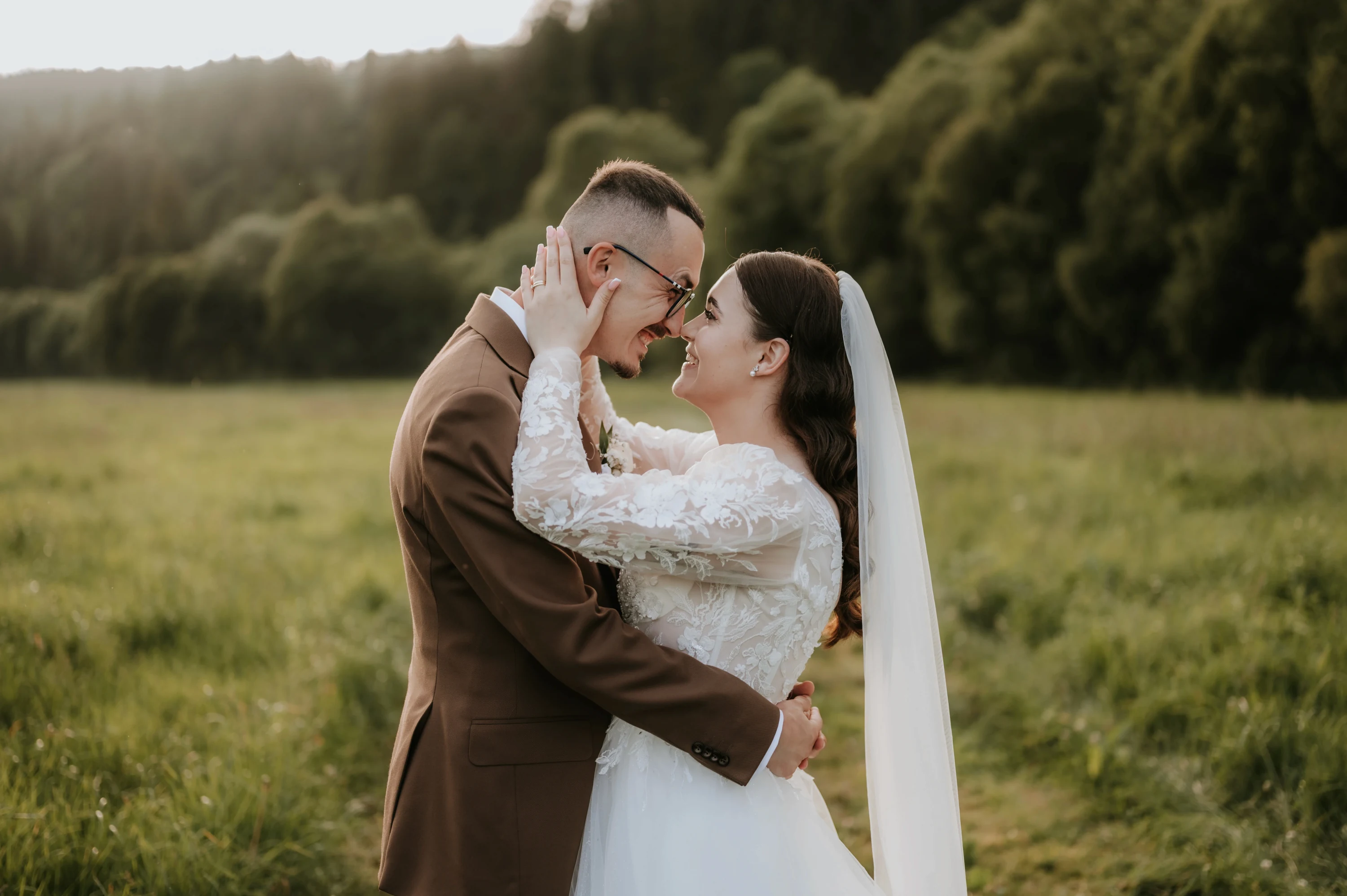 Nevesta so závojom sa dotýka tváre ženícha v hnedom obleku na lúke pred lesom.
