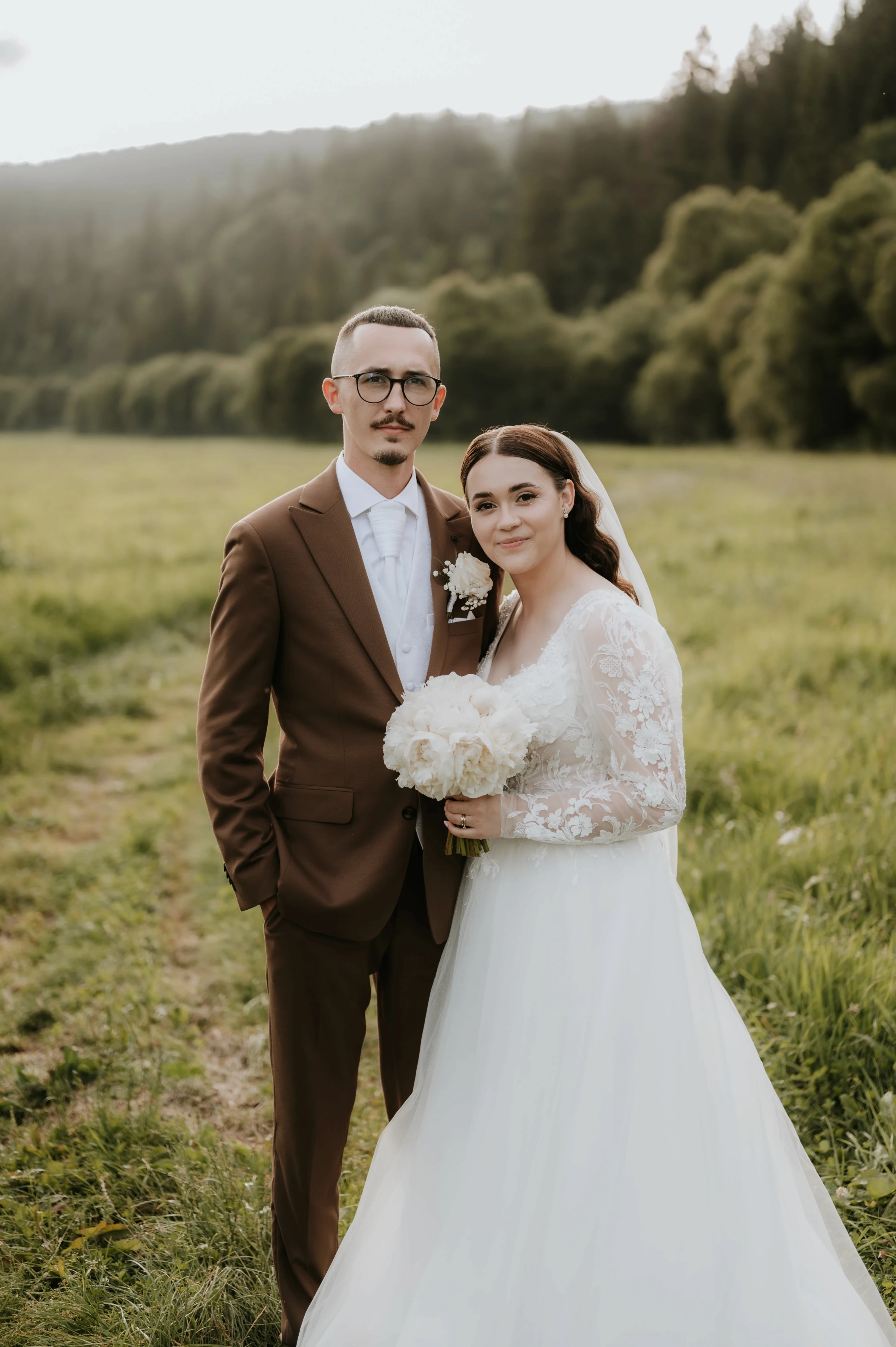 Pár pózuje na lúke pred horami, nevesta s bielou kytičkou v rukách.