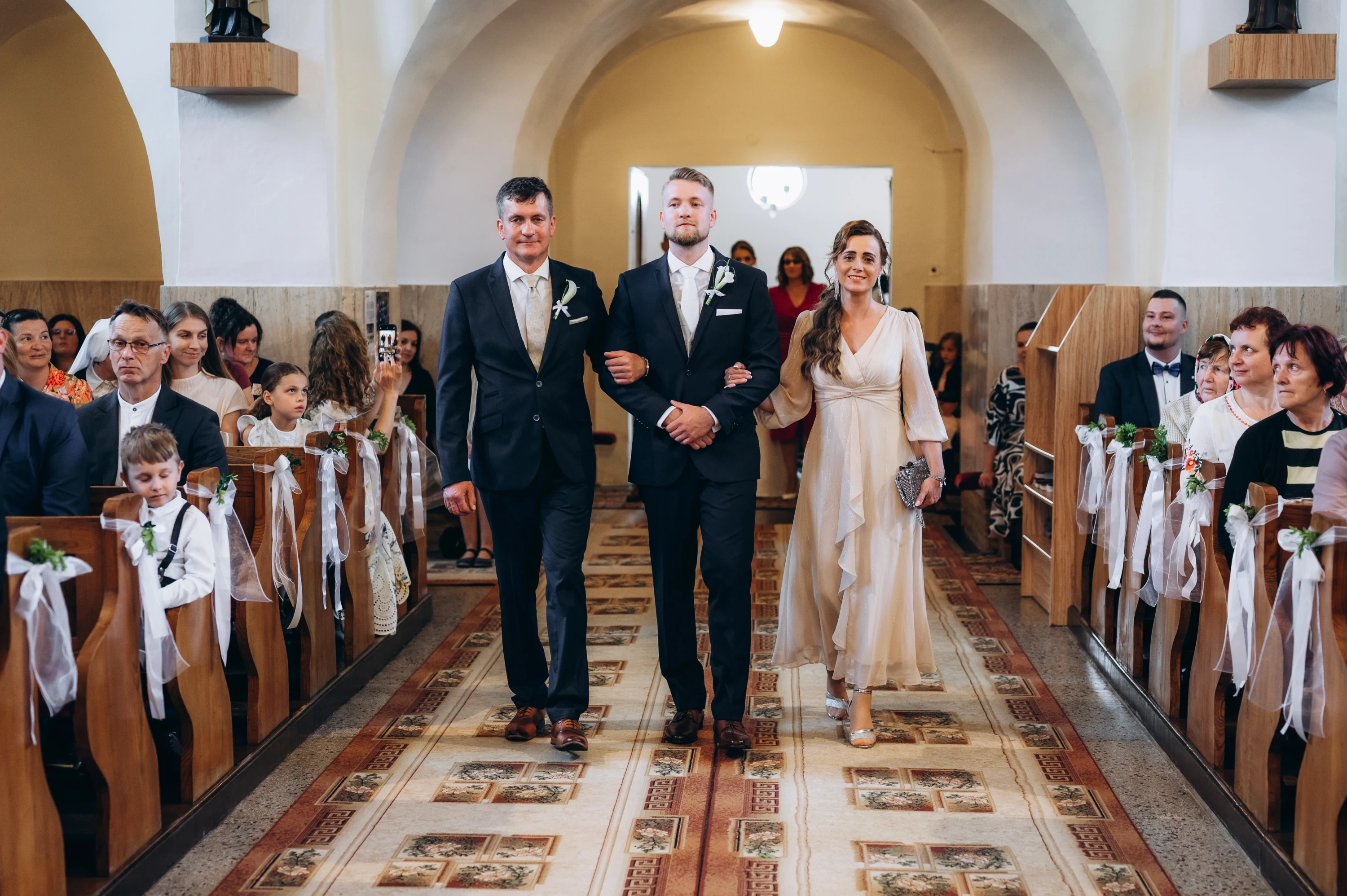 Ženích kráča kostolnou uličkou medzi starším mužom a ženou v šampanských šatách.