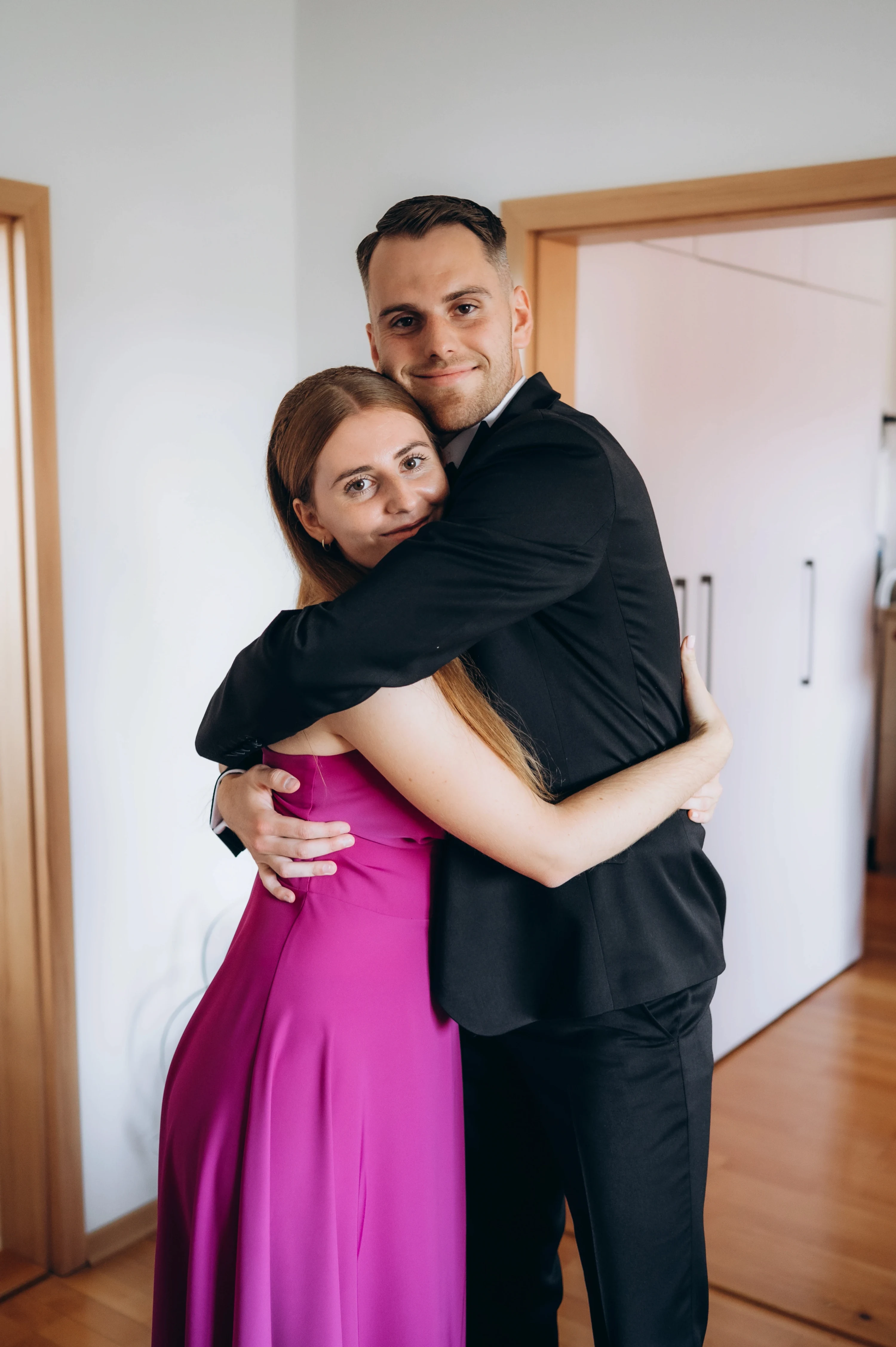 Muž v čiernom obleku pevne objíma ženu v purpurových šatách vo svetlej izbe.
