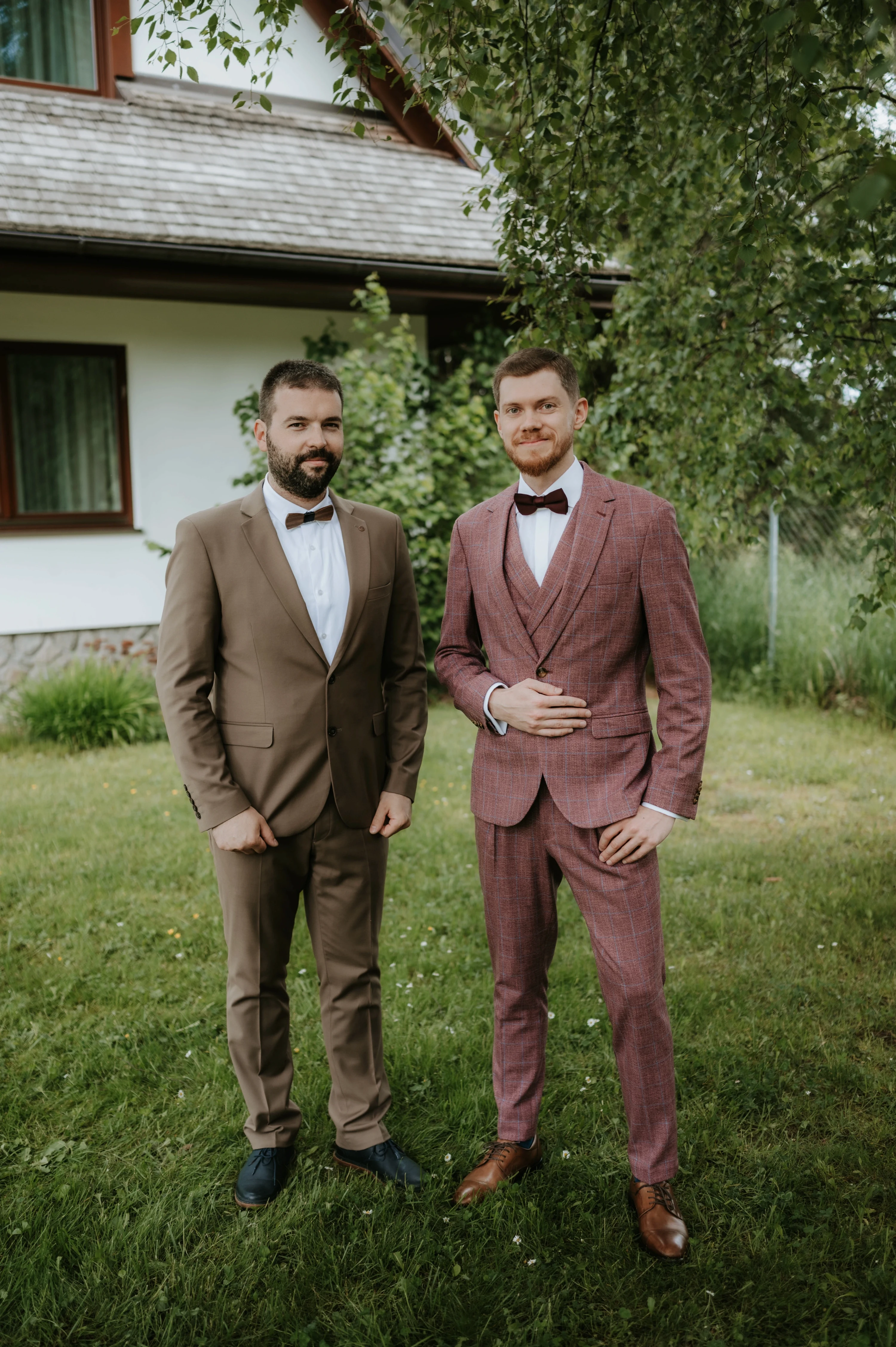 Dvaja muži v spoločenských odevoch sa smejú pred domom.