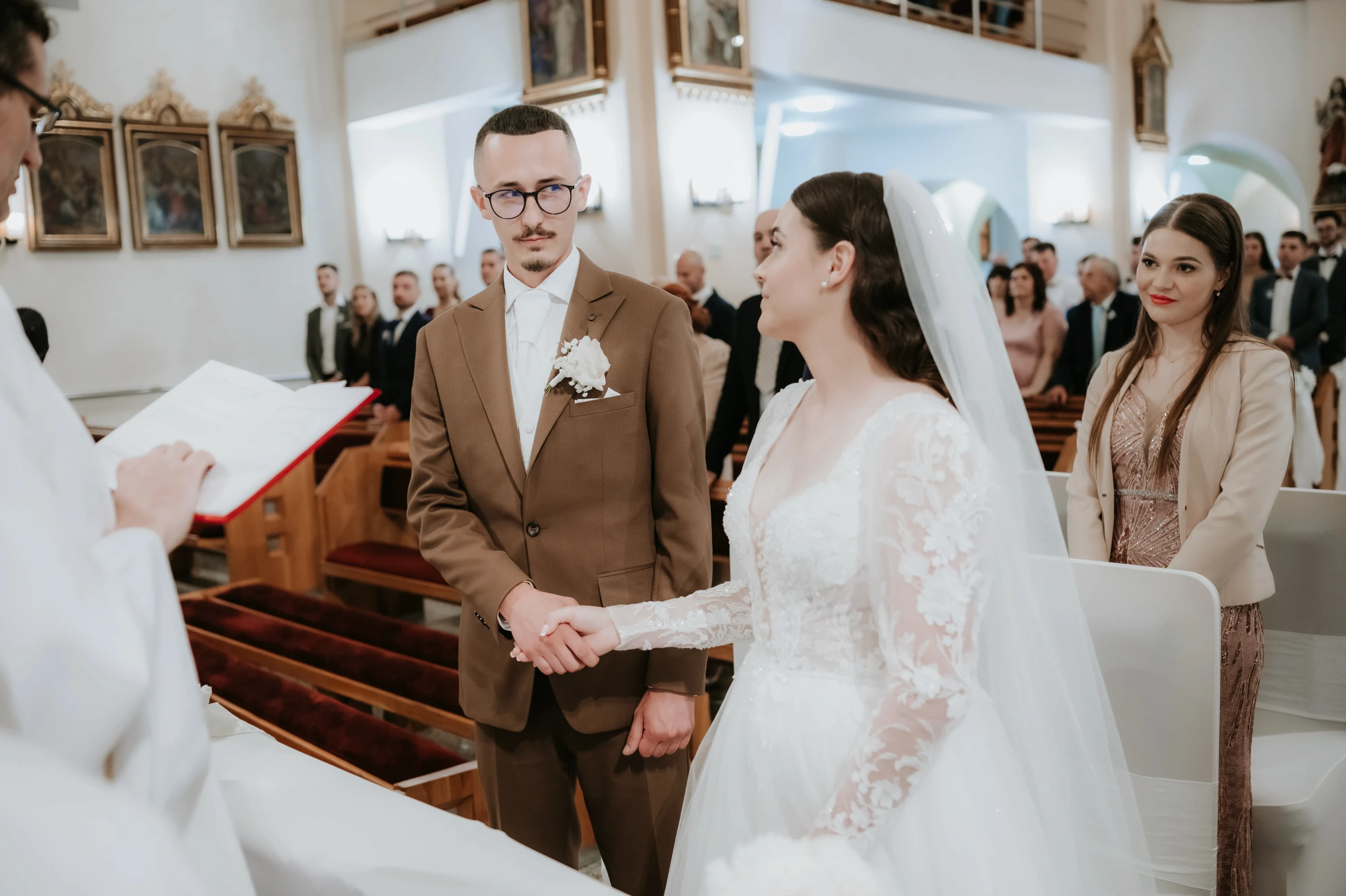 Ženích v hnedom obleku a nevesta v čipkových šatách si podávajú ruky pred kňazom.