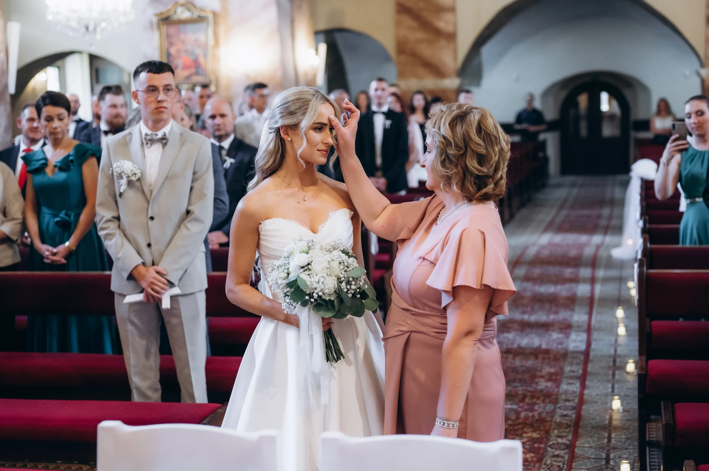 Žena v ružových šatách upravuje neveste vlasy v uličke kostola pred hosťami.