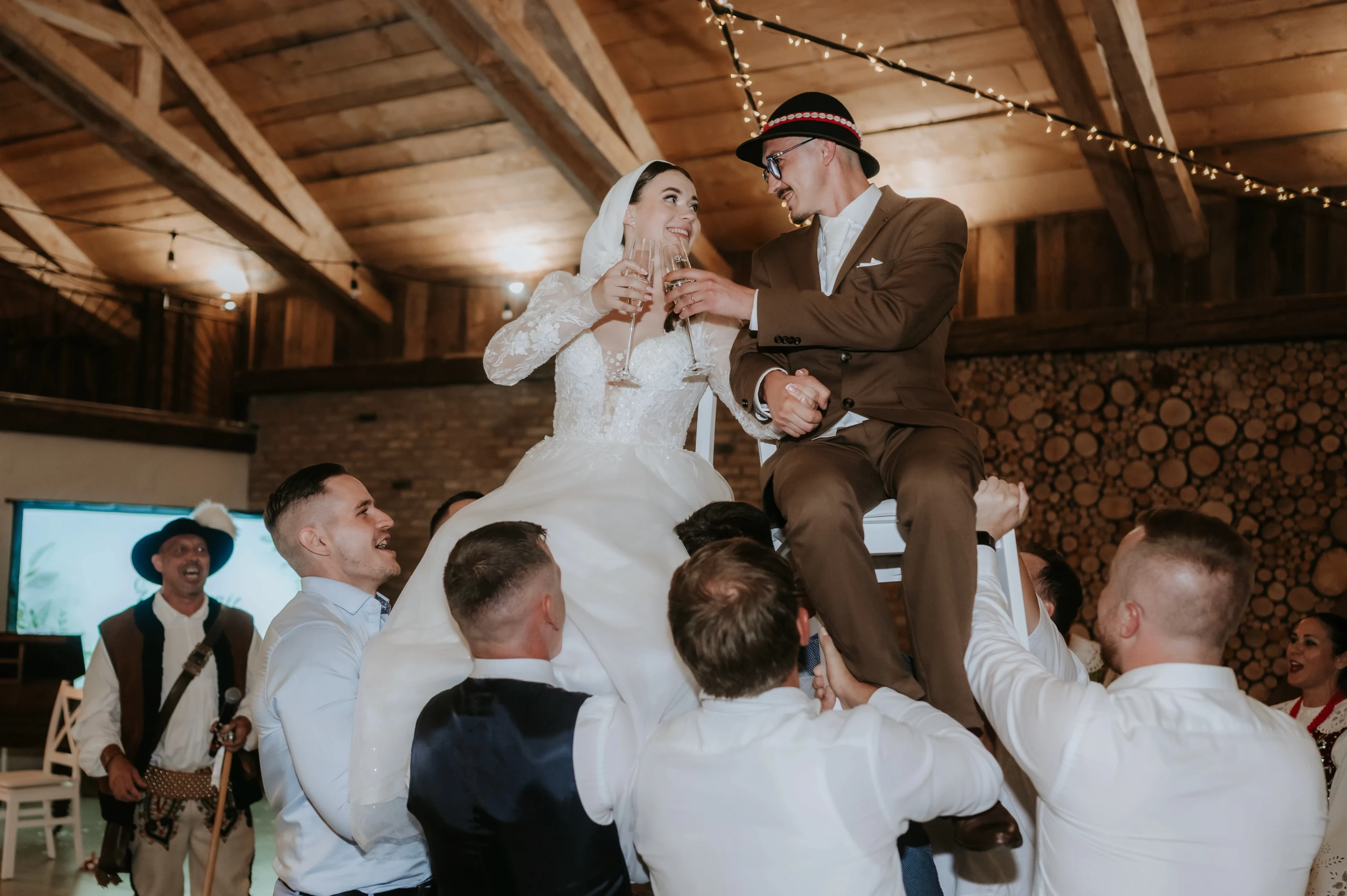 Hostia dvíhajú novomanželov na stoličkách v drevenej sále, pár si štrngá pohármi.