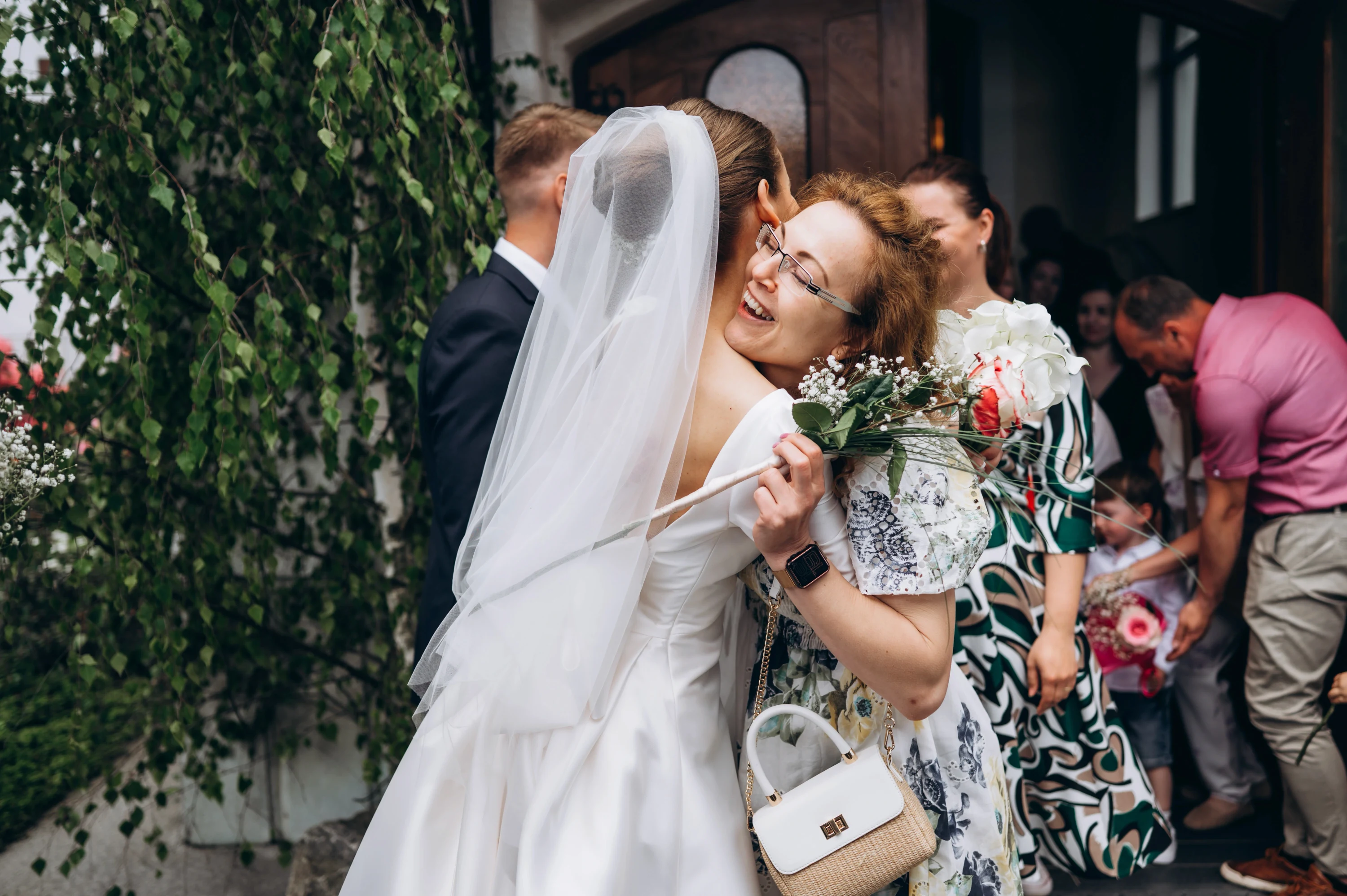 Nevesta v závoji objíma gratulantku s kyticou pri stene obrastenej zeleňou.