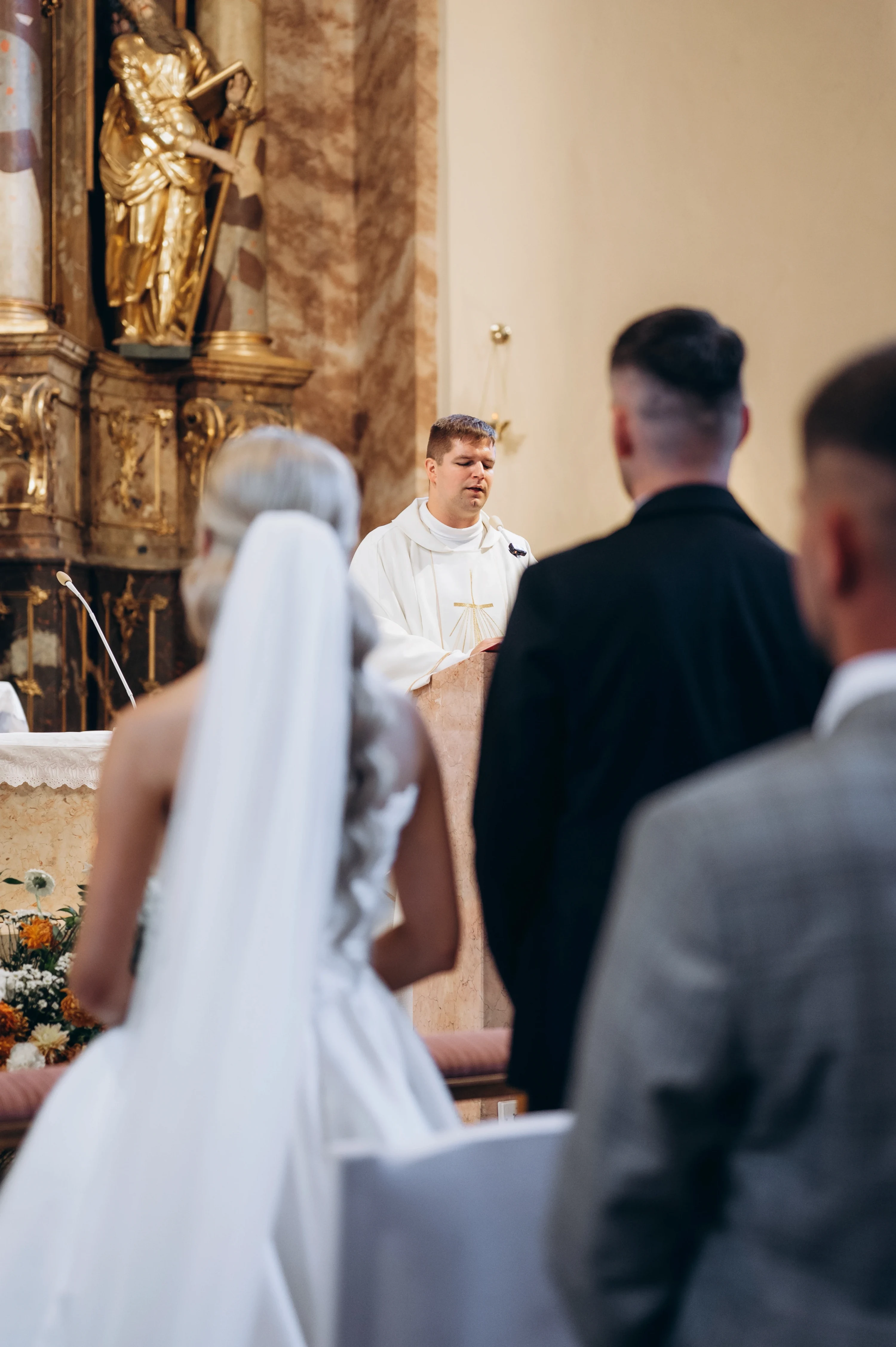 Kňaz hovorí pri oltári, v popredí sú chrbty nevesty a svadobčanov v oblekoch.