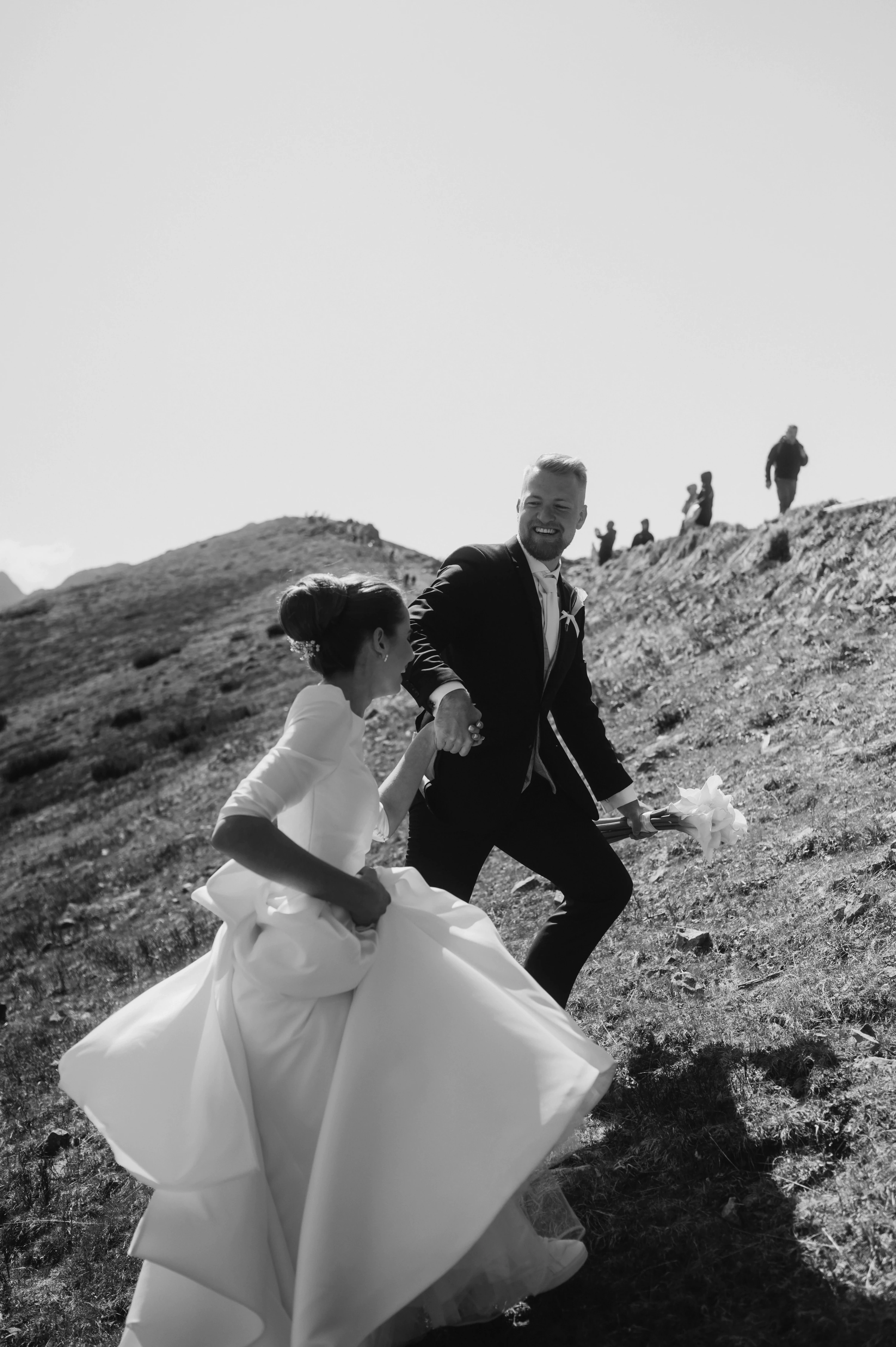 Rozosmiaty ženích vedie nevestu dolu trávnatým svahom na čiernobielej fotografii.