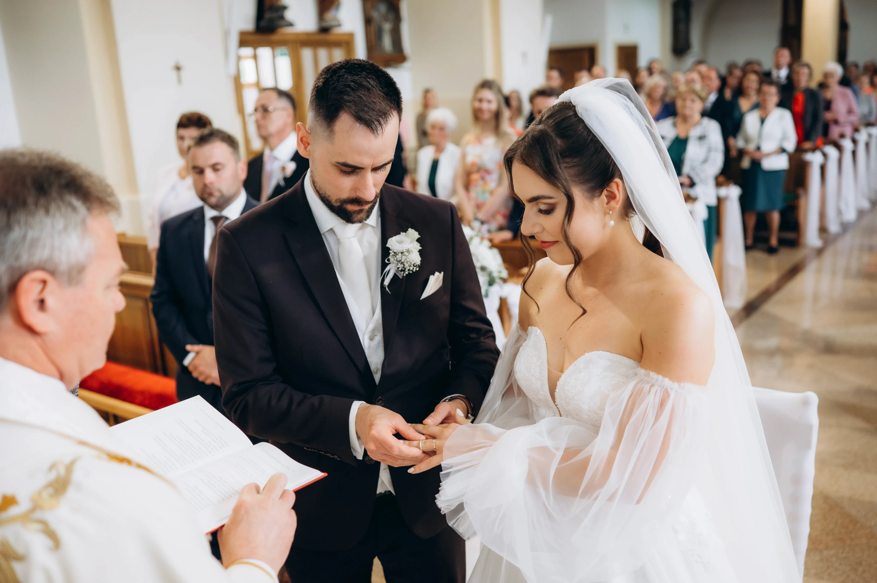 Ženích navlieka neveste obrúčku, v popredí kňaz číta z knihy počas obradu.