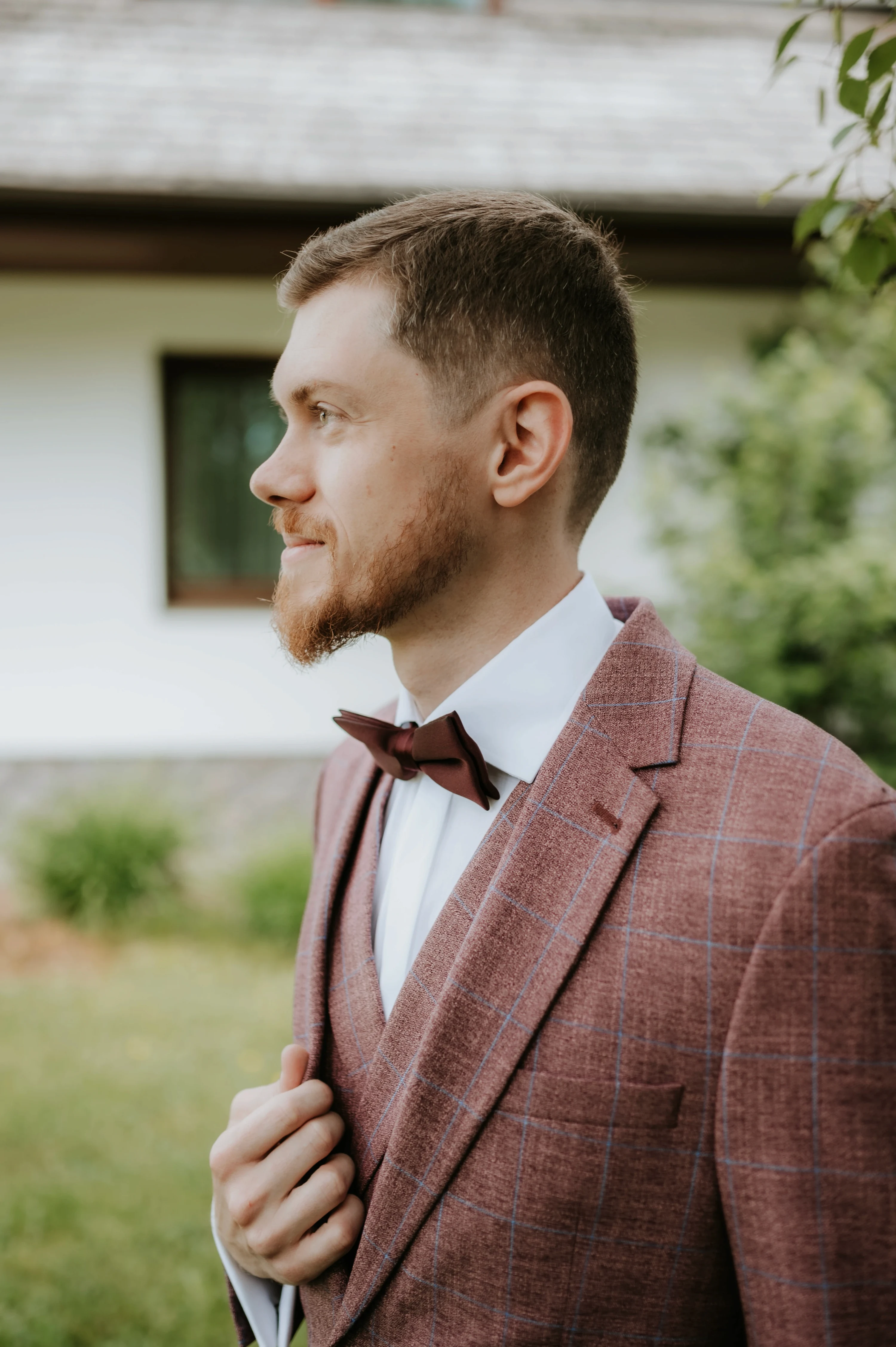 Ženích v károvanom vínovom saku s motýlikom stojí pred domom a hľadí do diaľky.
