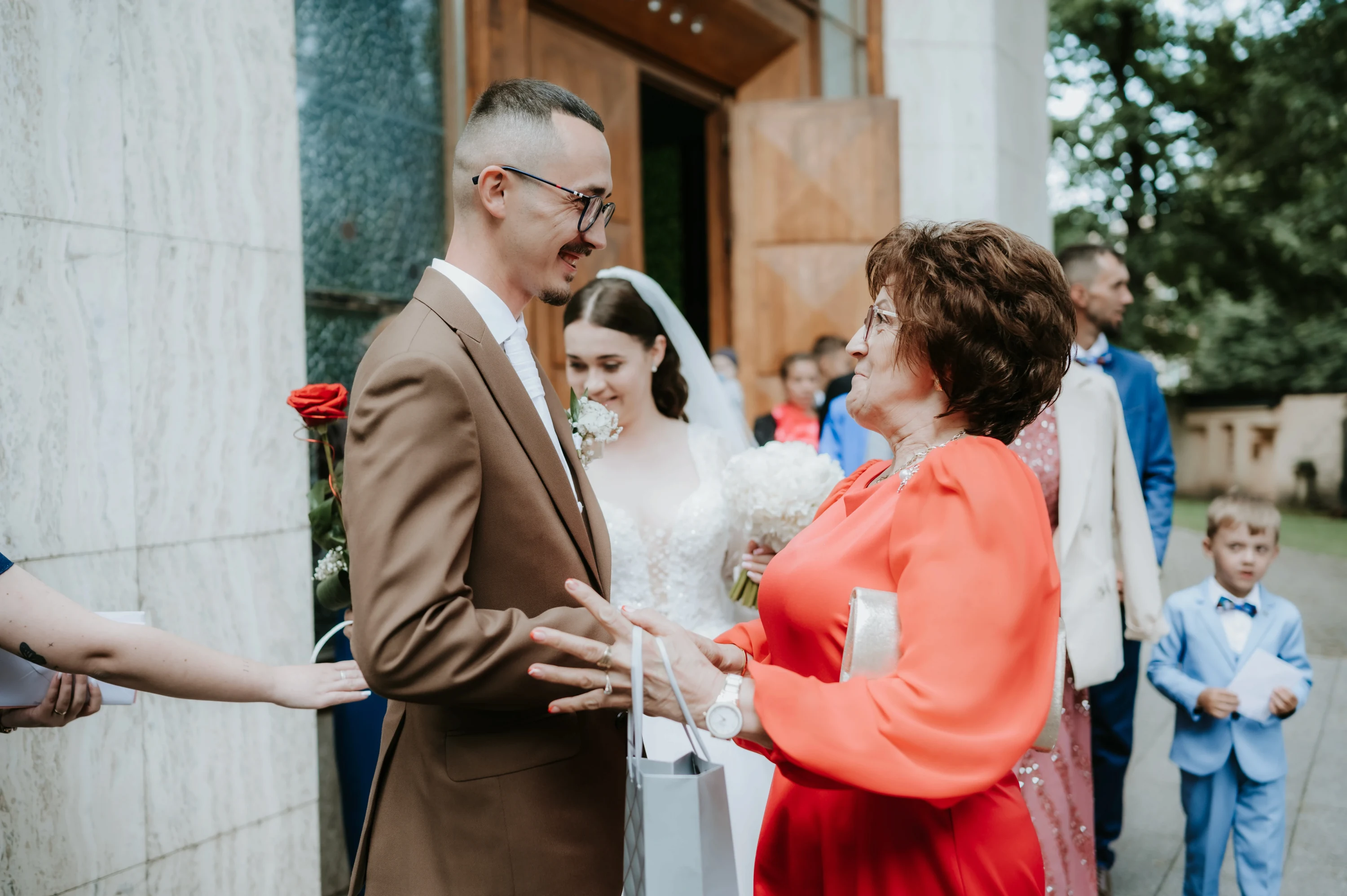 Ženích v hnedom obleku prijíma gratuláciu od ženy v oranžových šatách pred kostolom.
