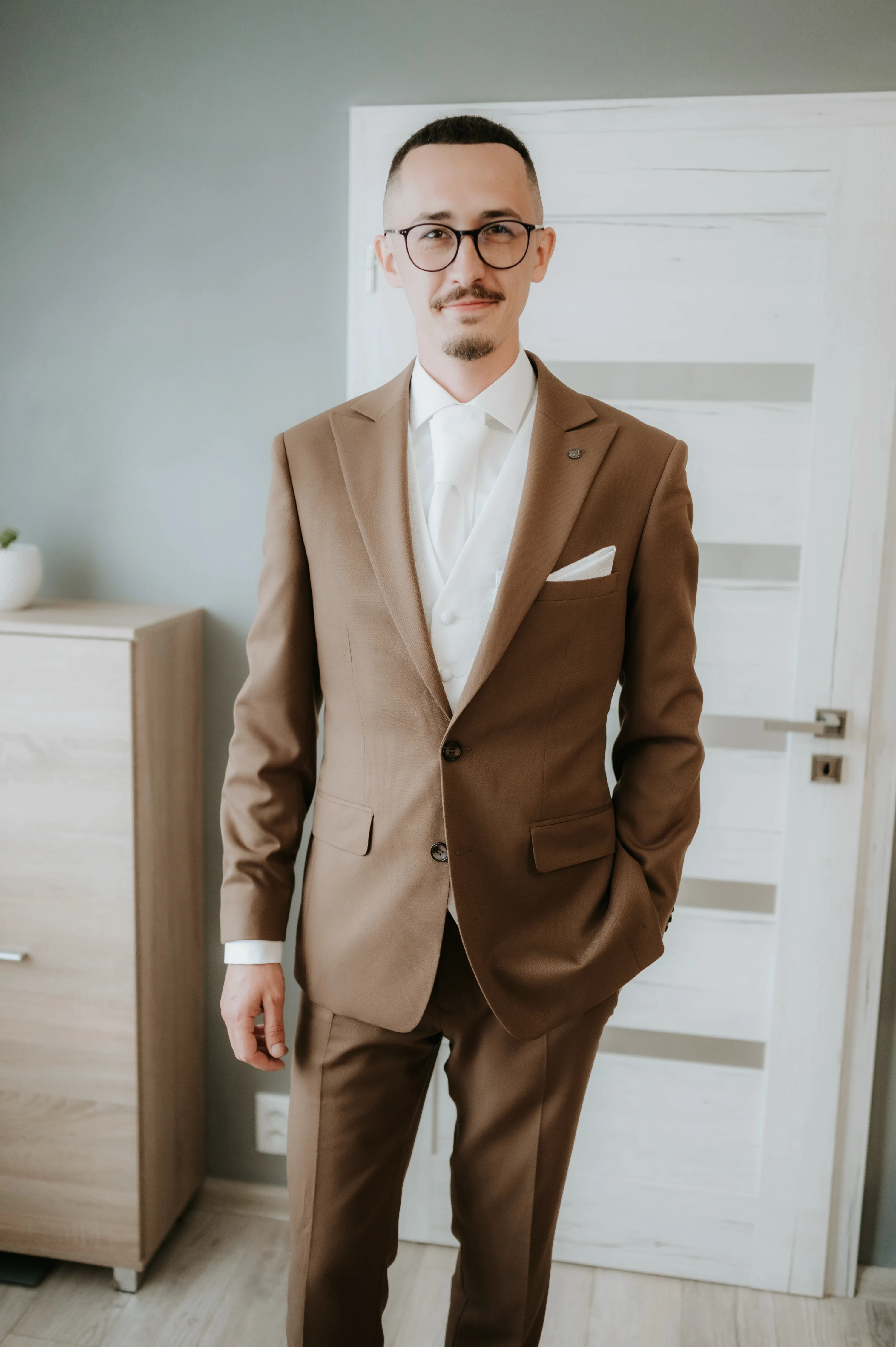 Ženích v hnedom obleku a bielej veste stojí pri dverách izby pred odchodom.