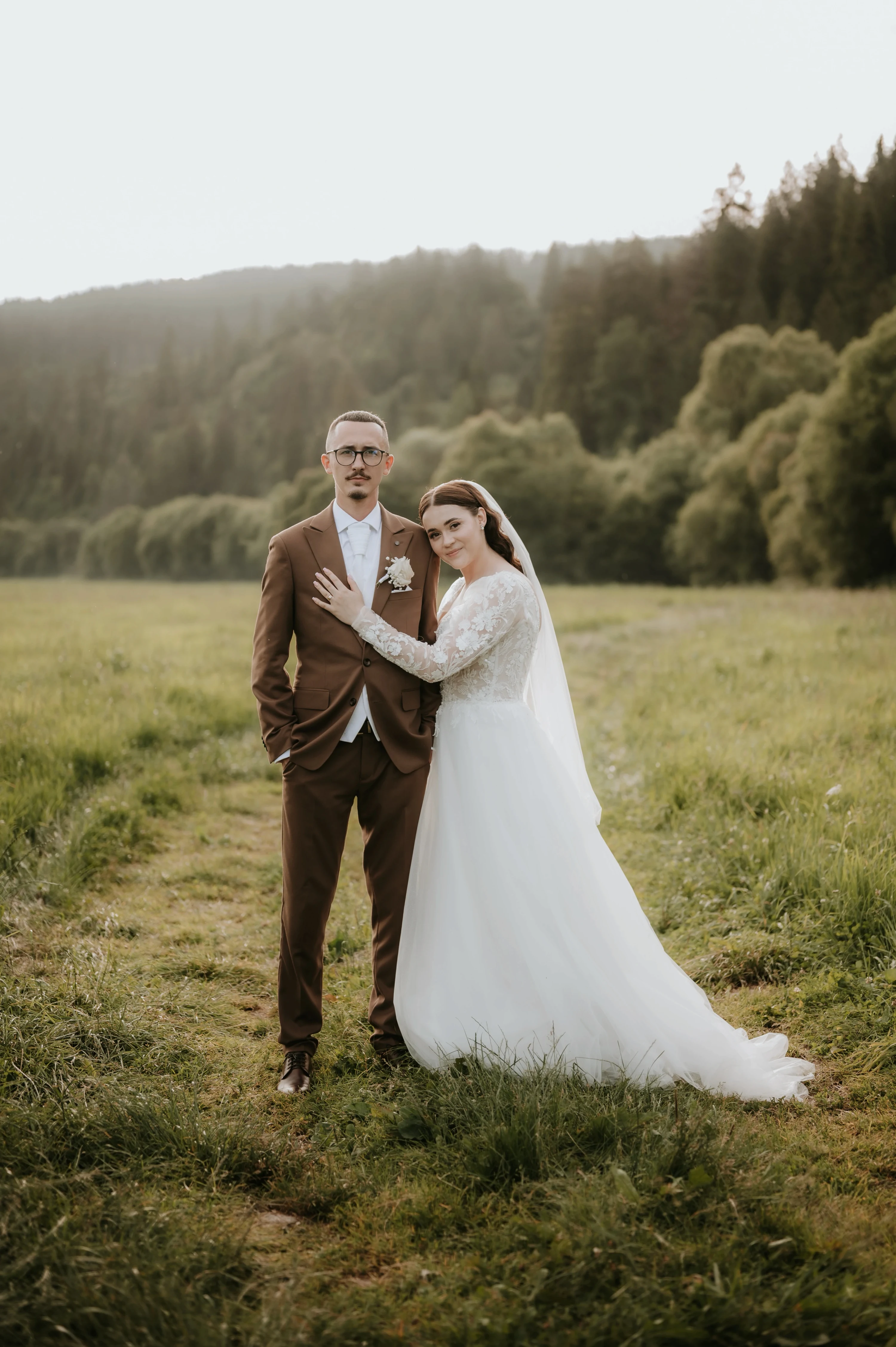 Novomanželia stoja v objatí na lúke pred zalesnenými kopcami.