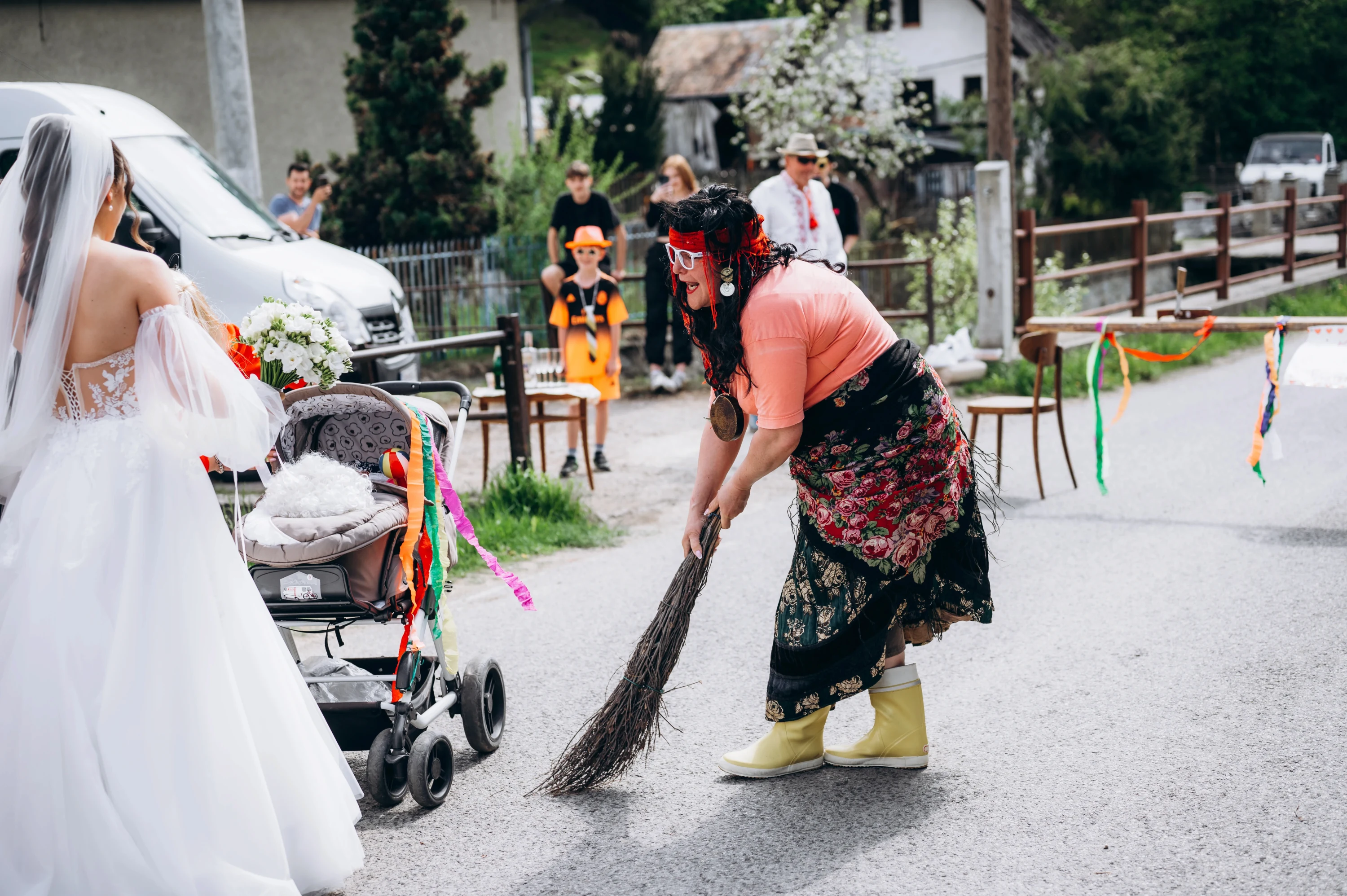 Žena v ružovej košeli metie cestu pred nevestou v bielych svadobných šatách.