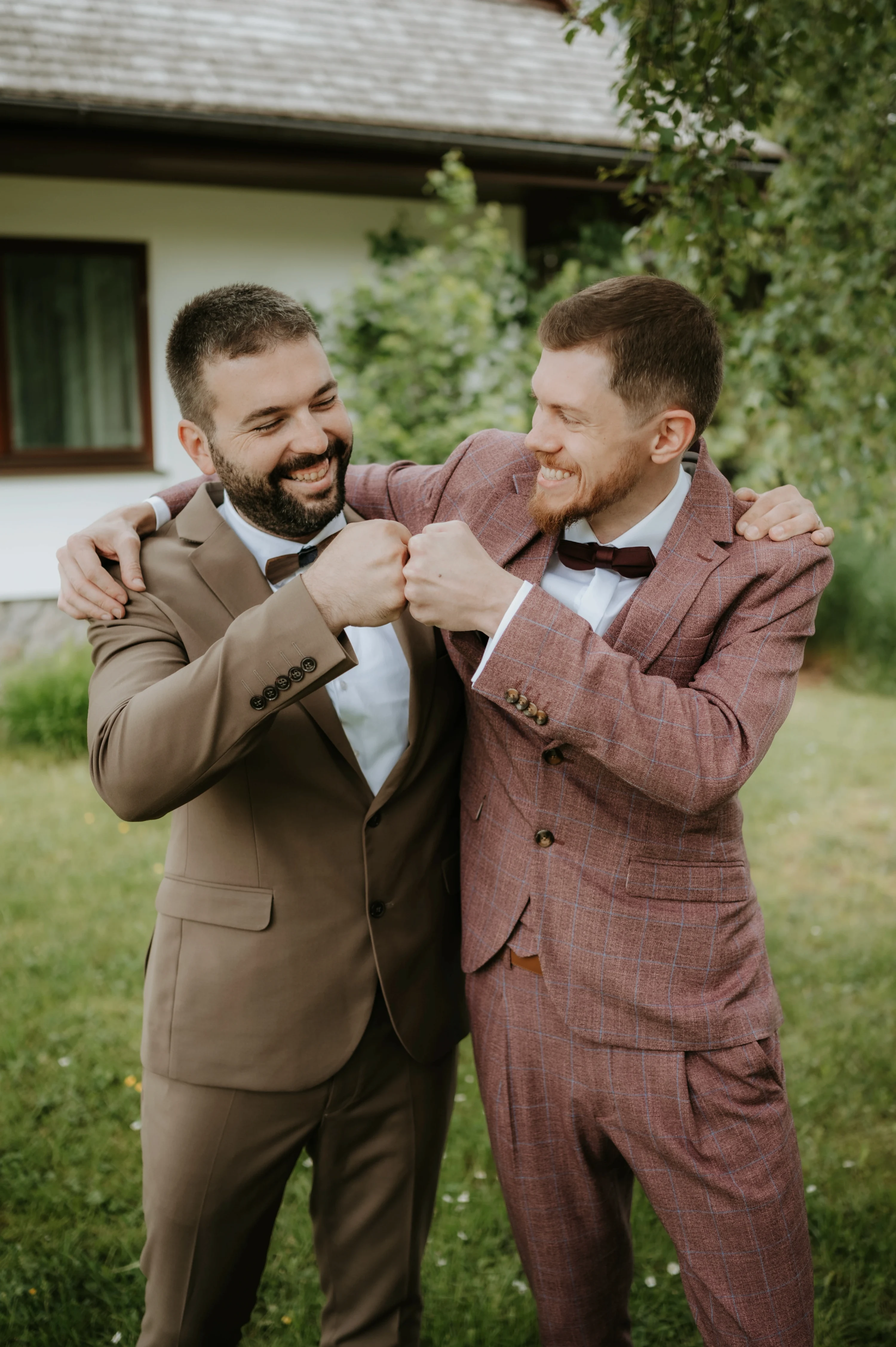 Dvaja muži v oblekoch sa smejú a ťukajú si päsťami pred domom.
