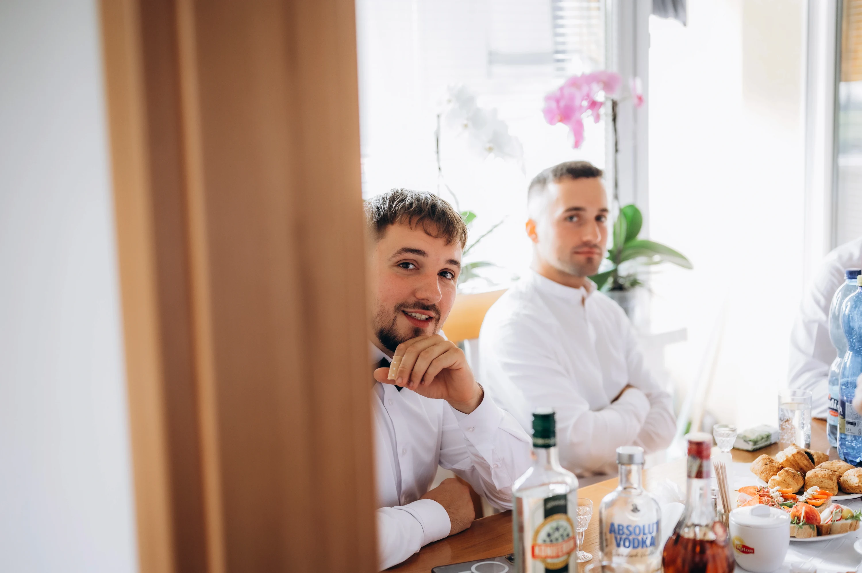 Dvaja muži v bielych košeliach sedia za stolom s fľašami a chlebíčkami počas príprav.