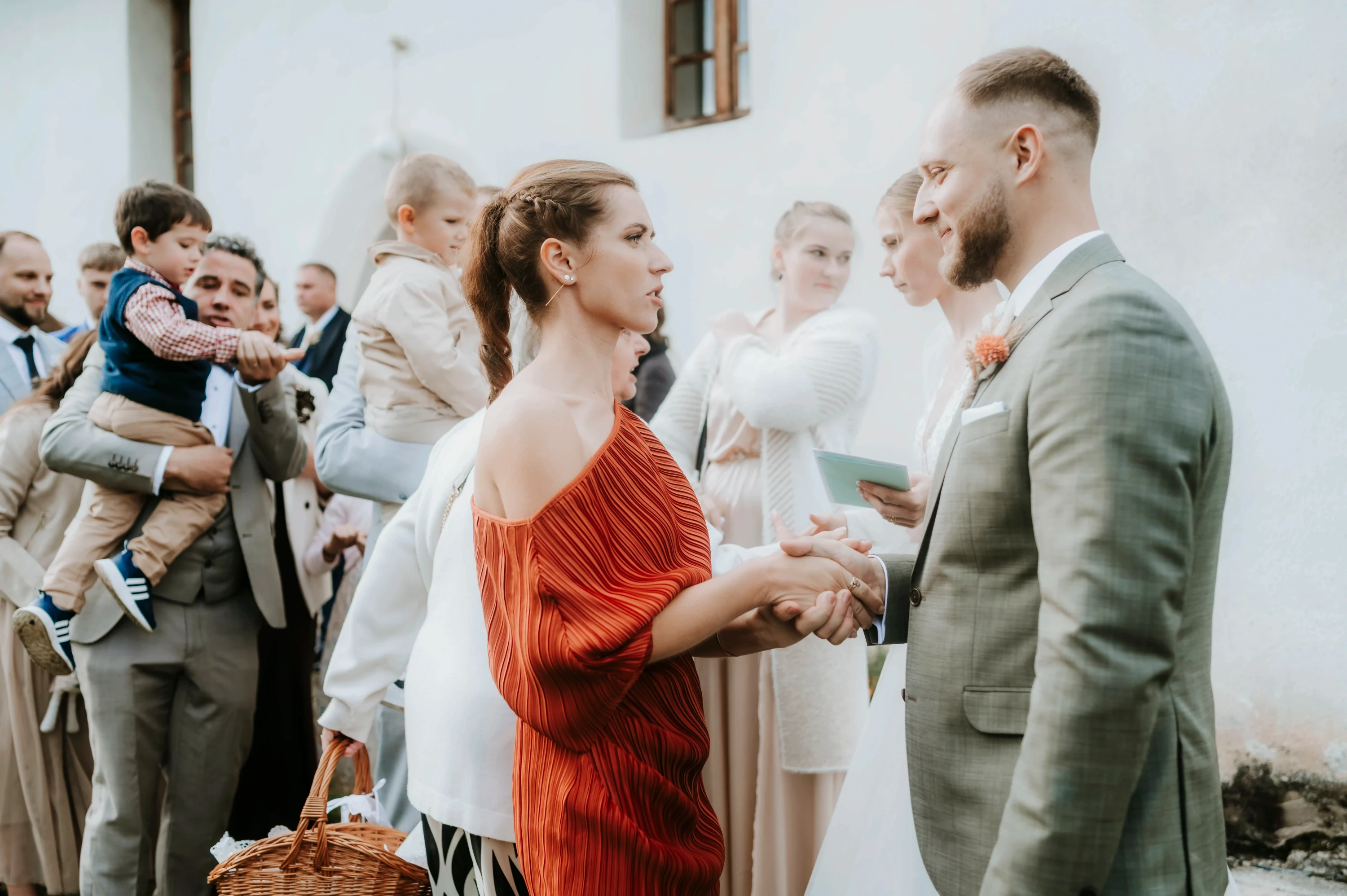 Ženích si podáva ruku s gratulantkou v oranžových šatách pred bielym kostolom.