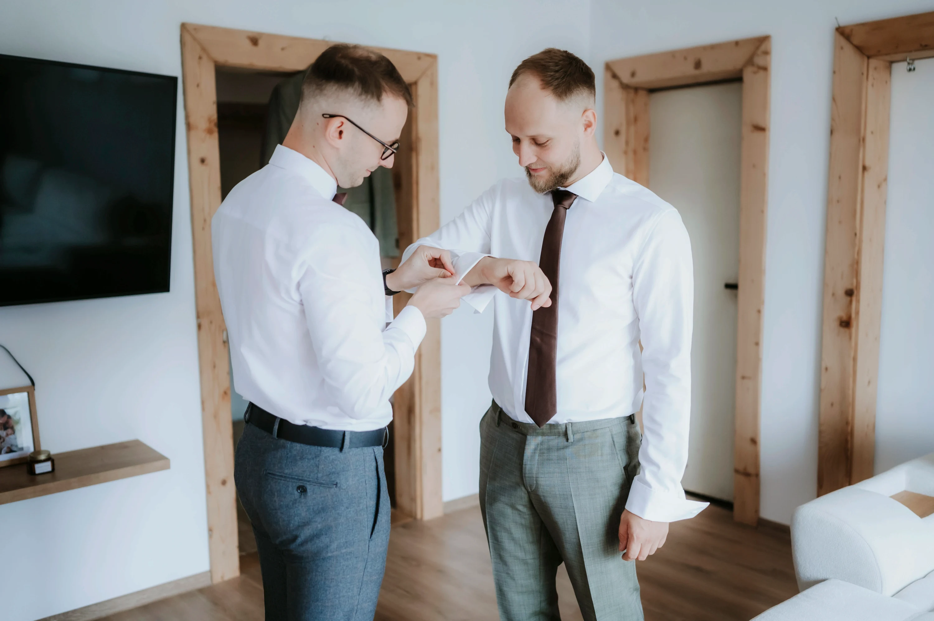 Muž v bielej košeli pomáha ženíchovi so zapínaním manžety pred odchodom.
